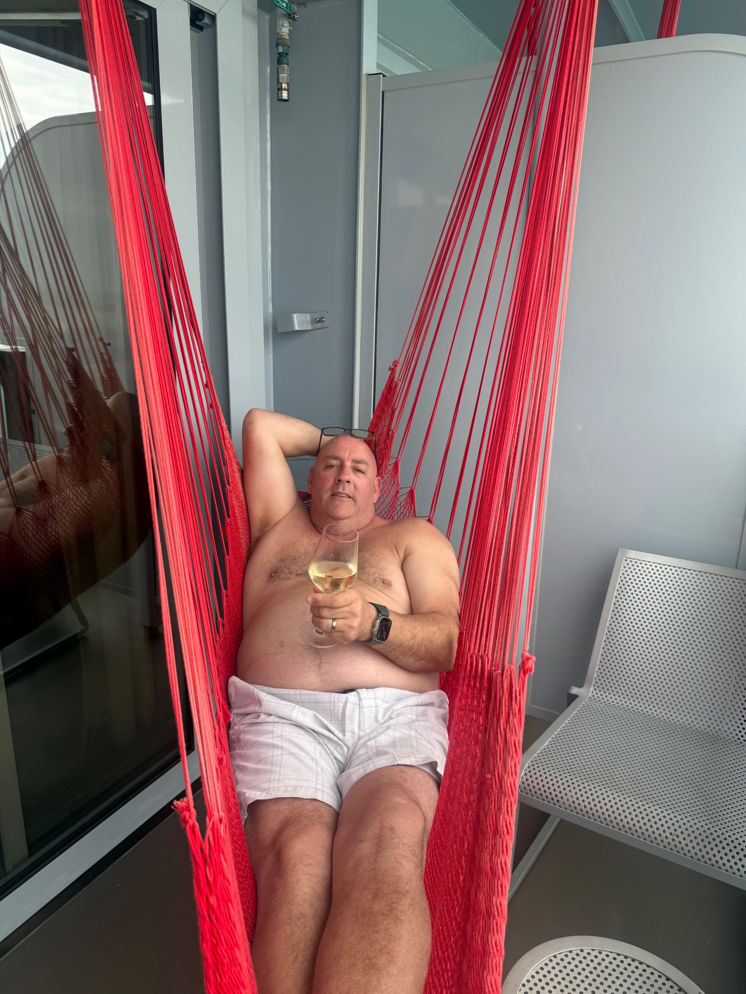 Man relaxing in red hammock on ship balcony holding glass of white wine, shirtless in casual vacation setting