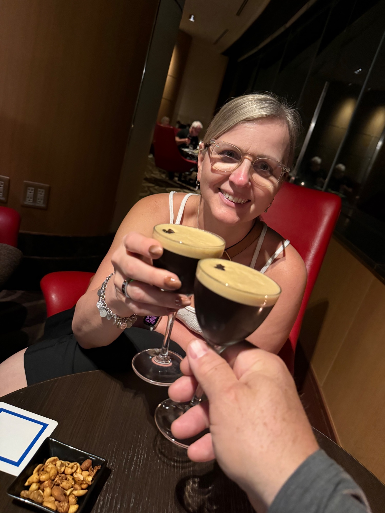 Woman holding two dark cocktails with wooden lids at a table with nuts and snacks visible