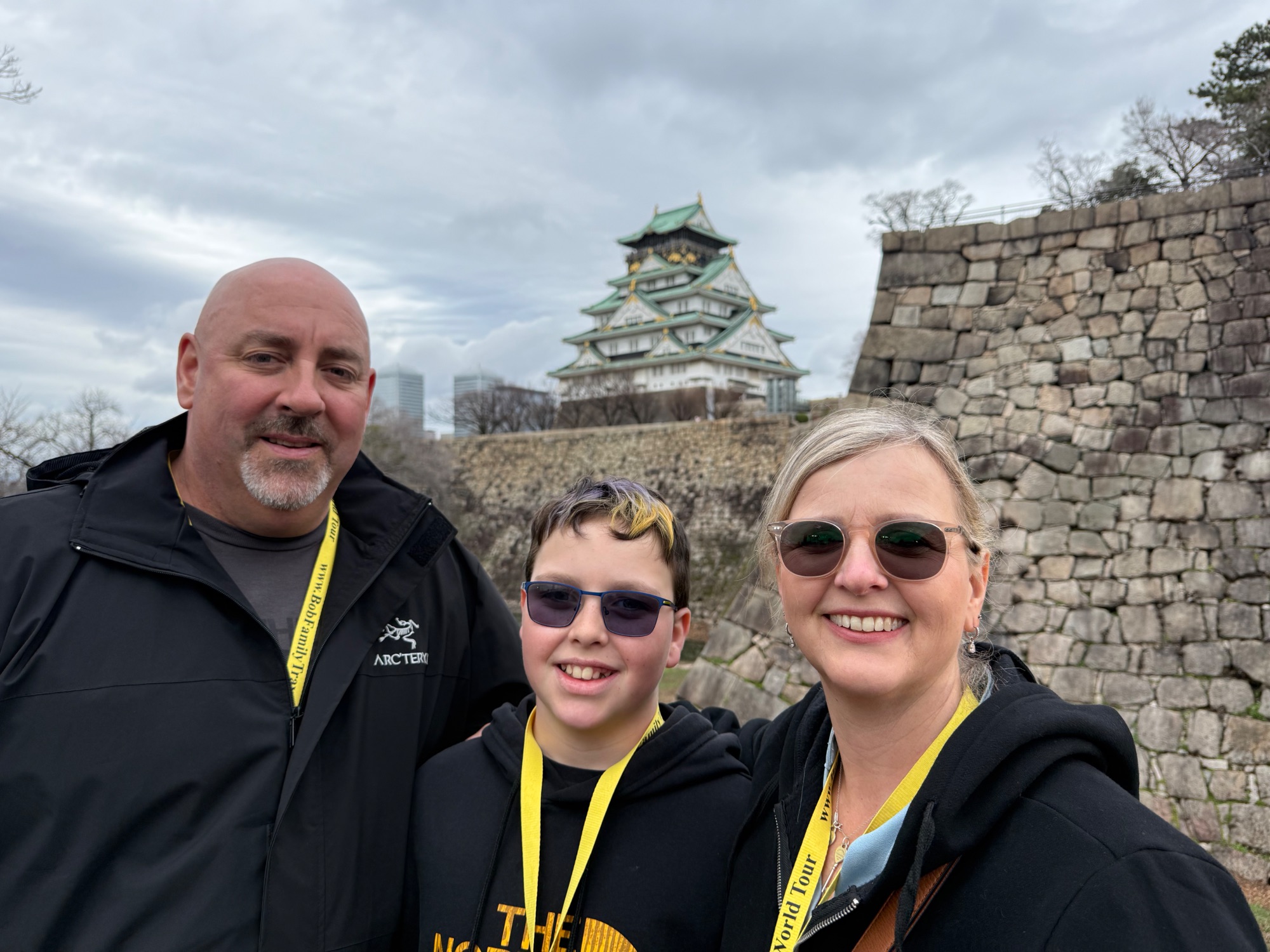 Family of three wearing yellow lanyards at Osaka Castle with traditional architecture visible behind them