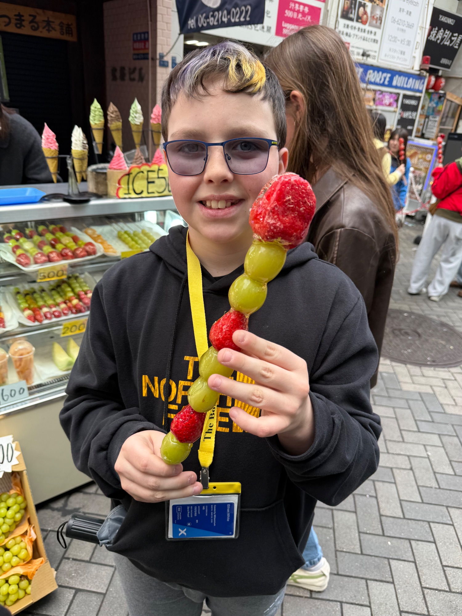 Young boy holding a colorful dango skewer with red and green fruit balls at a Japanese street market