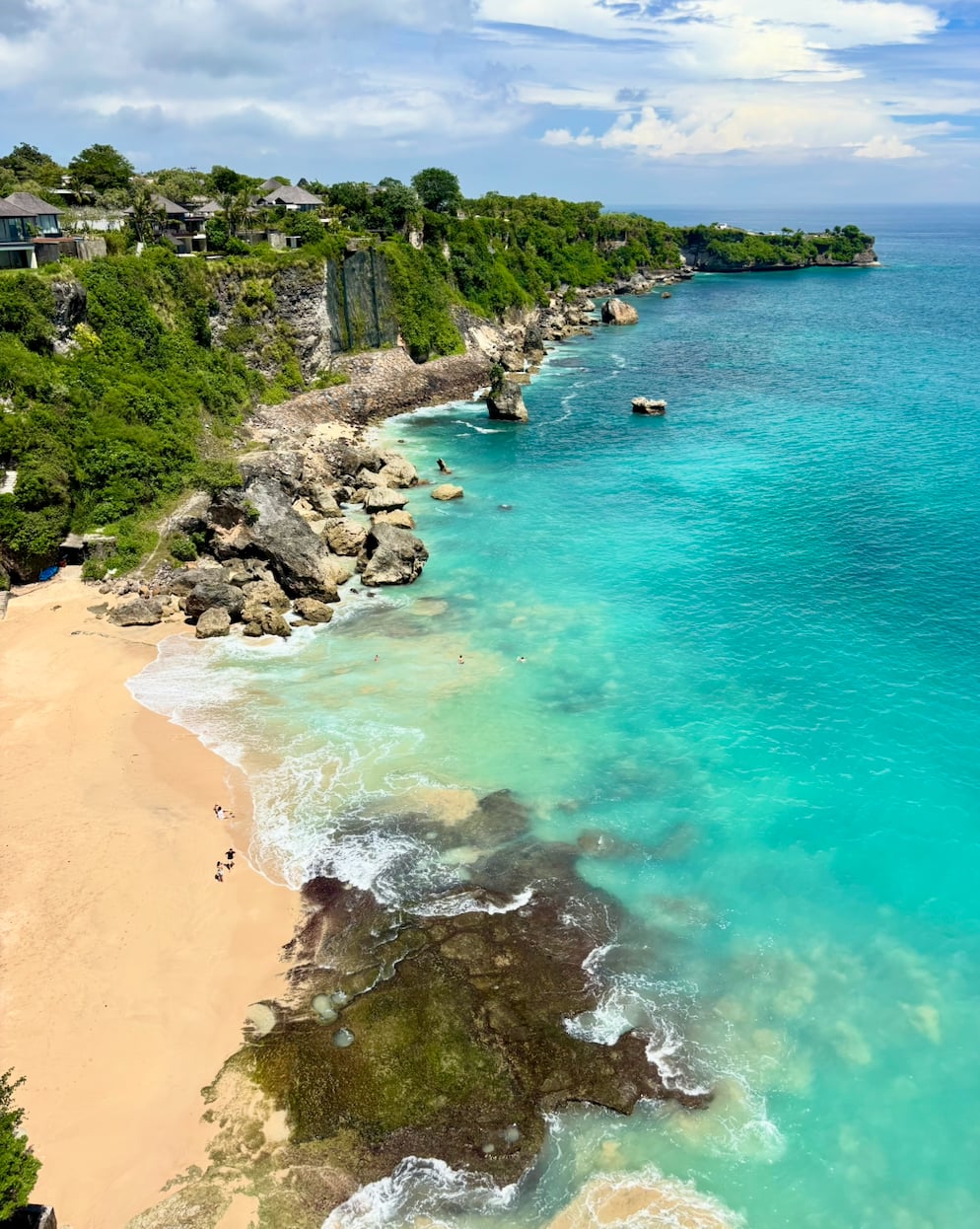 Aerial view of a turquoise cove with sandy beach, rocky cliffs, and traditional Balinese structures on the hillside