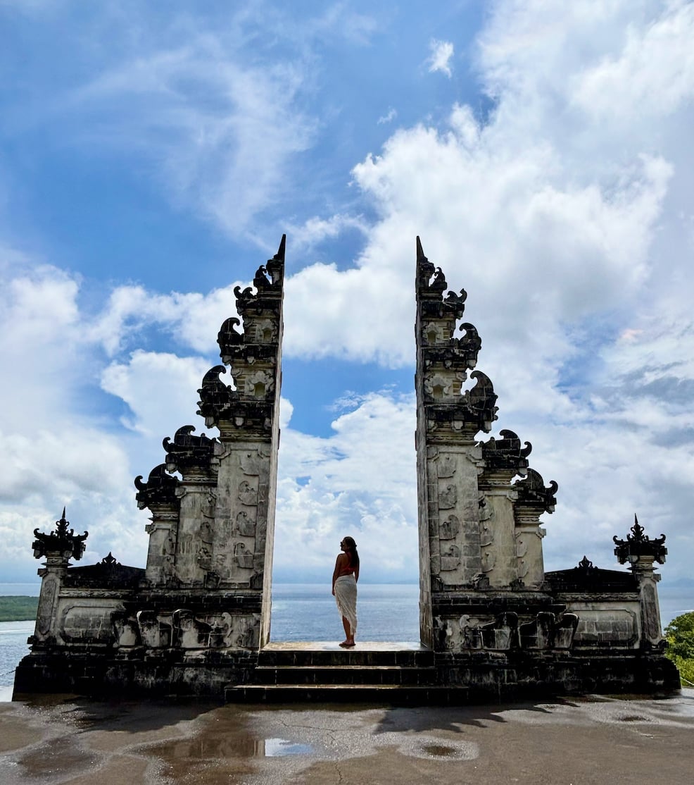 Julia Cianci stands between two tall ornate stone temple gates with ocean and sky in the background