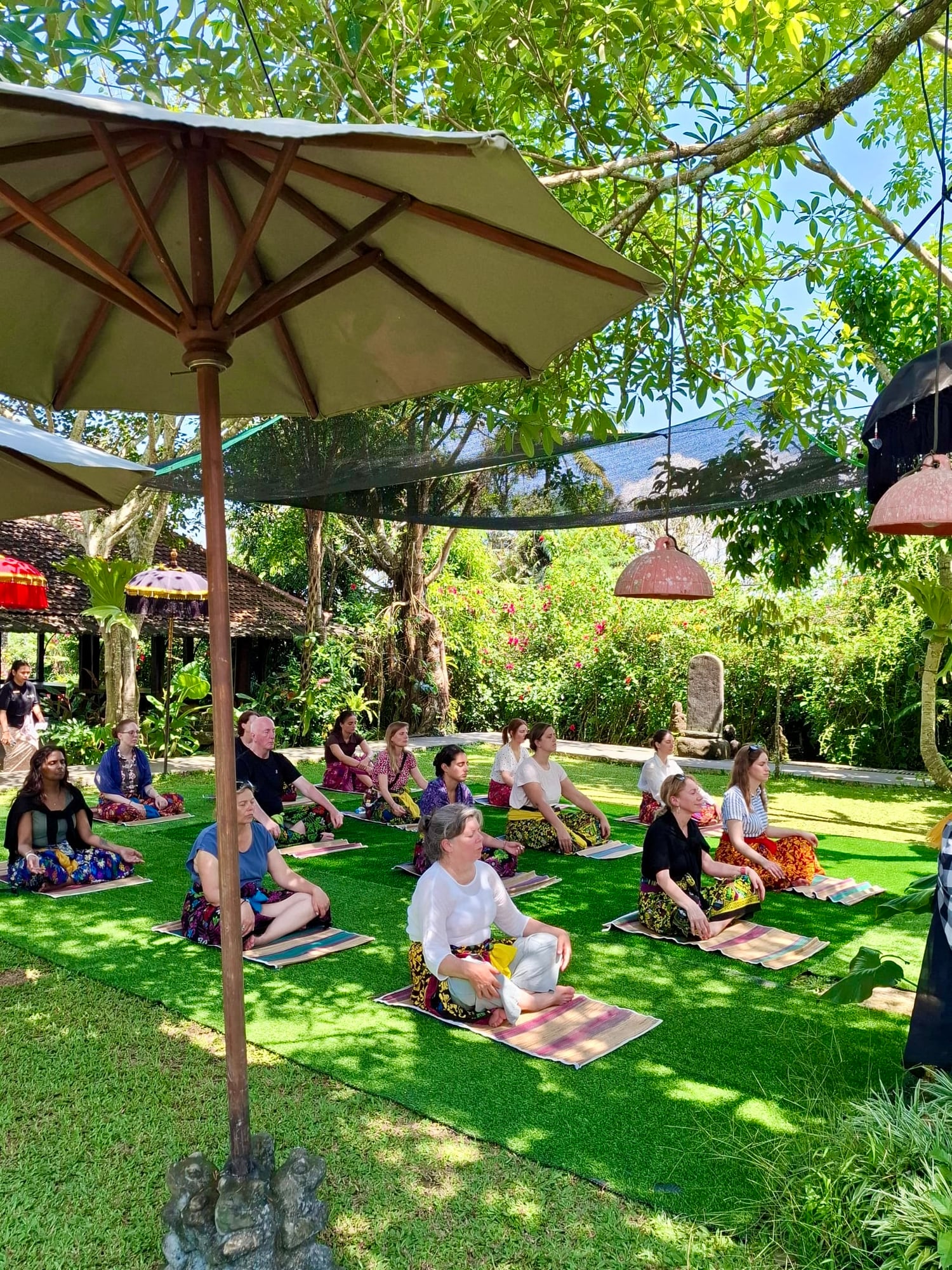 Outdoor yoga session on grass mats under umbrellas with participants in various poses among tropical gardens