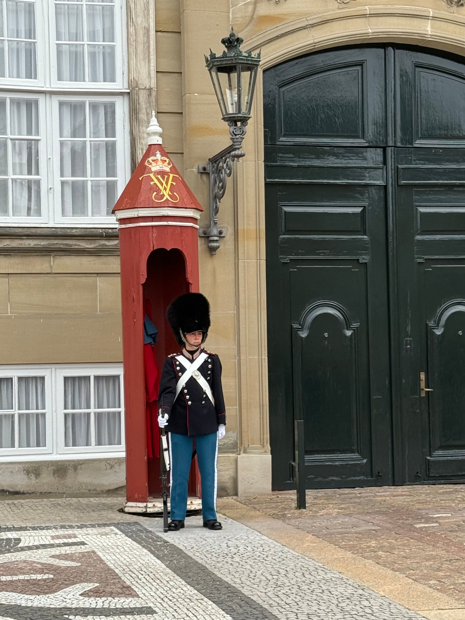 A uniformed royal guard in red jacket and bearskin hat stands at attention beside a red sentry box