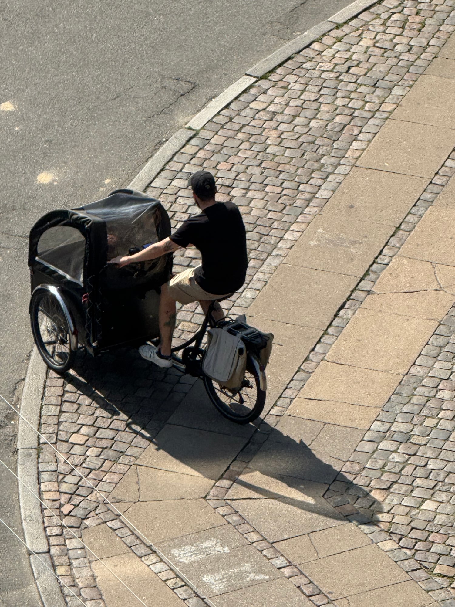 A person riding a cargo bike with a black canopy on a brick-paved street, viewed from above