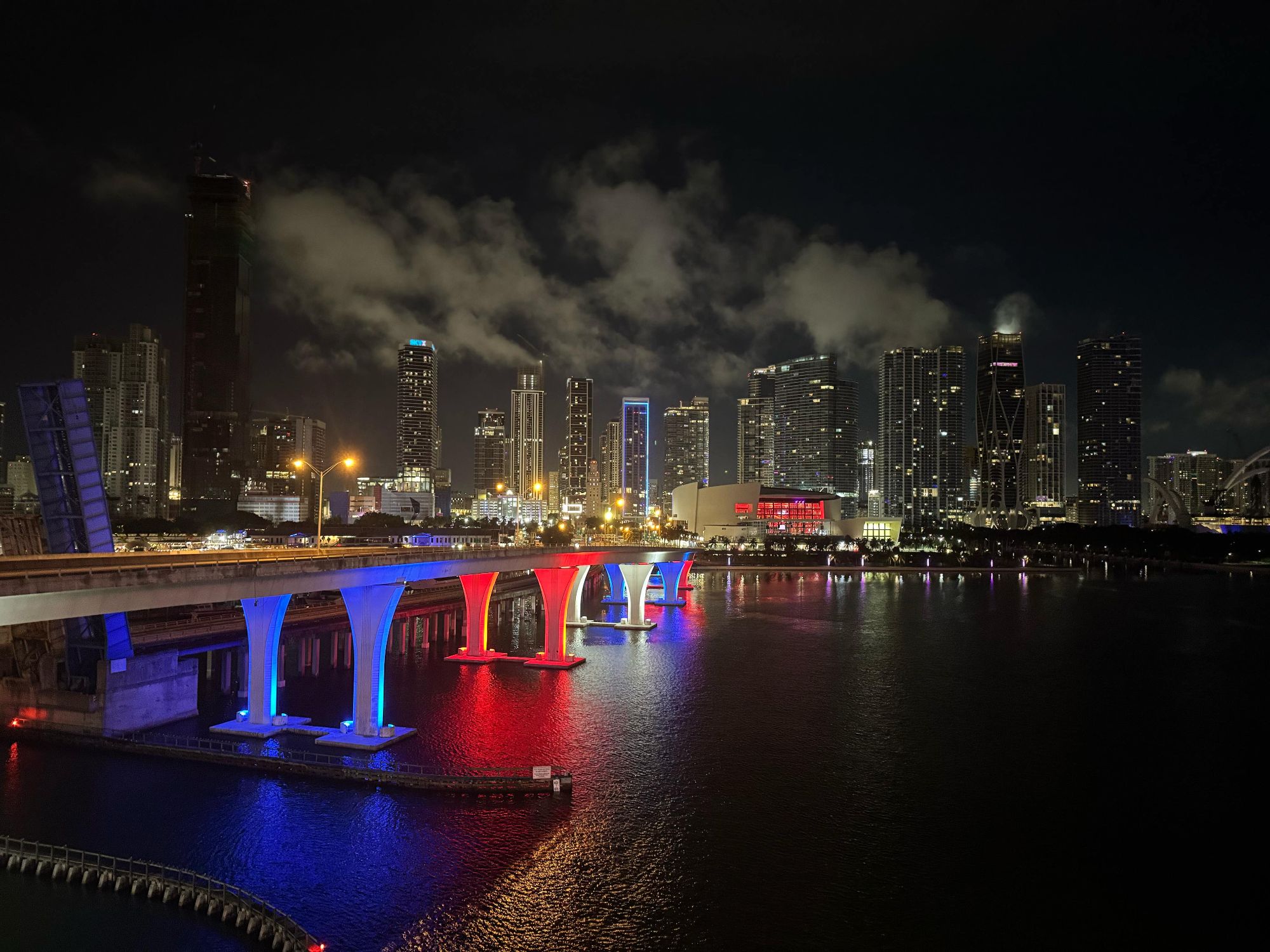 Miami's causeway bridge glows red, white, and blue against a luminous downtown skyline reflected in Biscayne Bay at night.