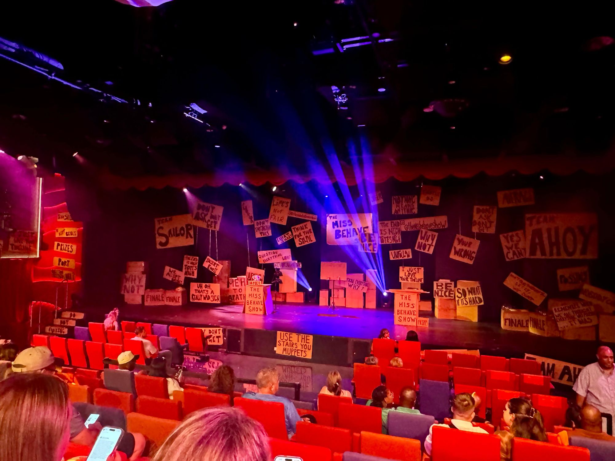 Audience awaits The Miss Behave Show inside a vibrant theater covered in humorous handwritten signs.