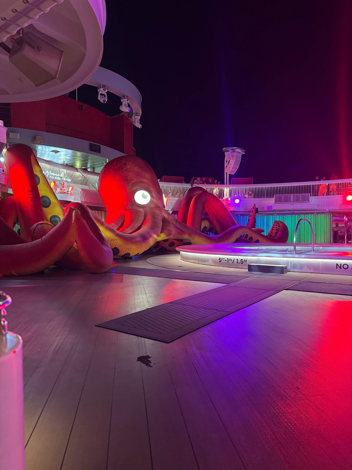 A massive inflatable octopus sprawls across a cruise ship pool deck under dramatic party lights at night.