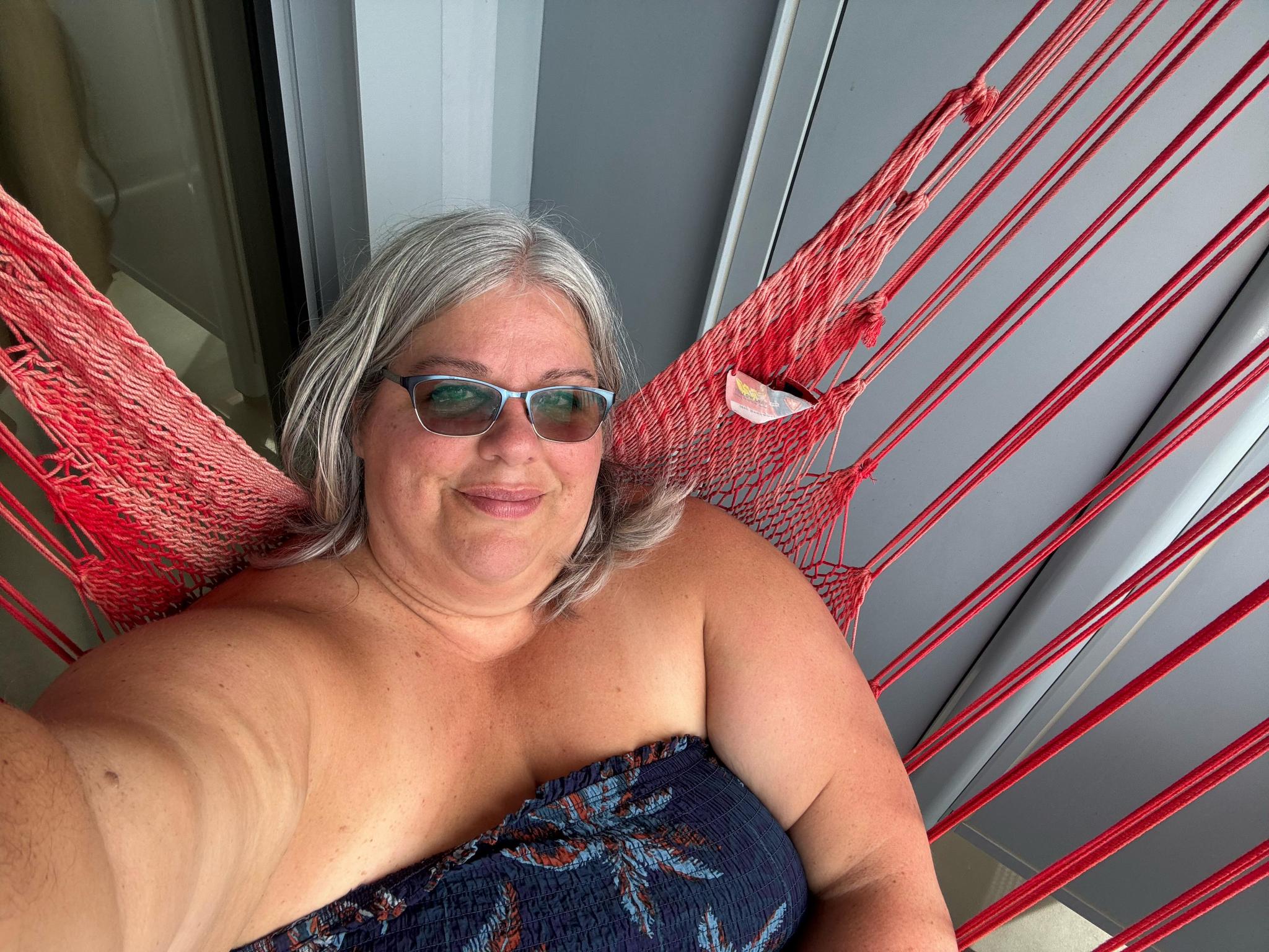 A woman relaxes in a vibrant red hammock on a covered balcony, smiling at the camera.