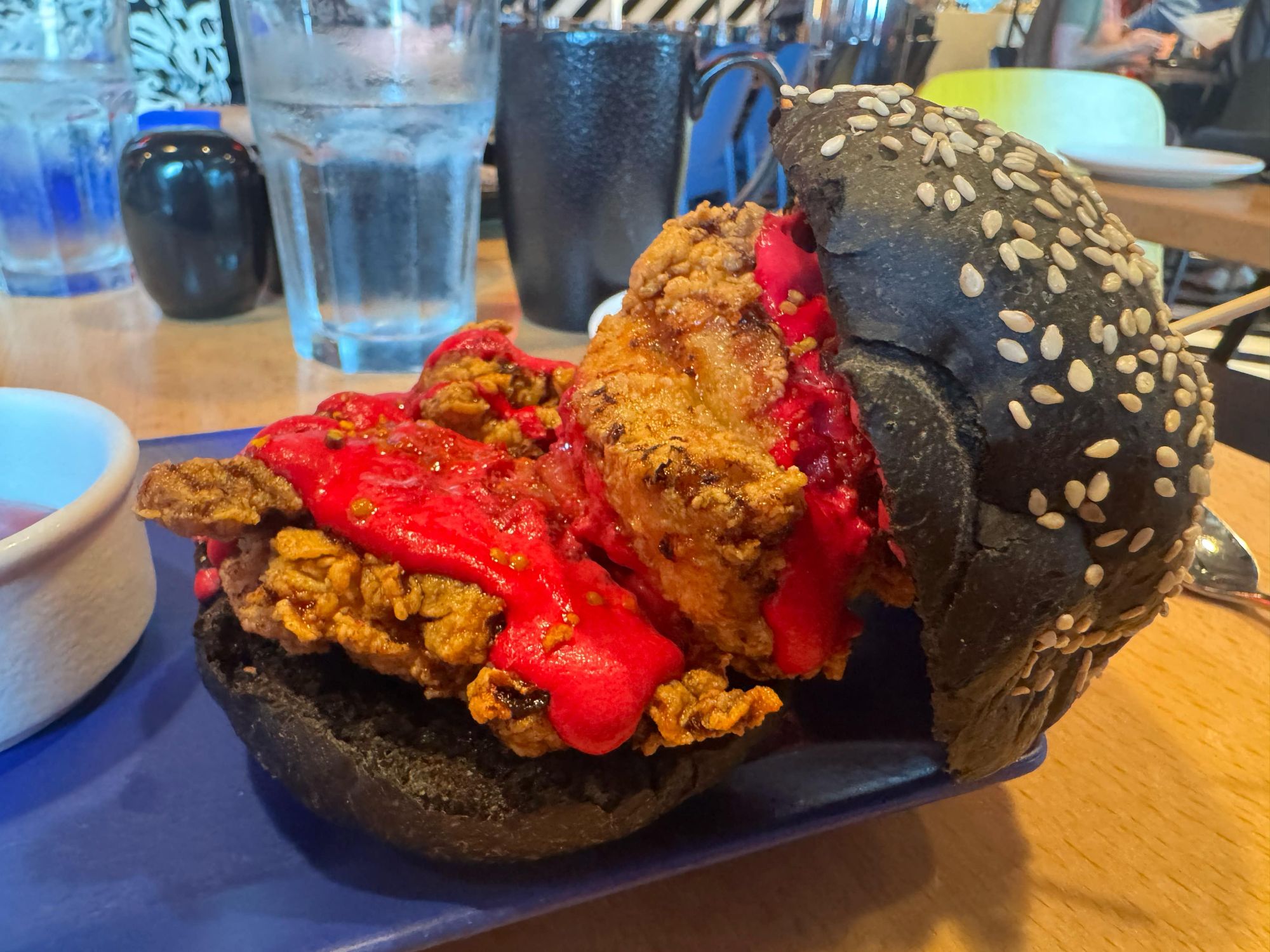 A dramatic black-bun fried chicken sandwich drenched in vivid red spicy sauce served at a restaurant.