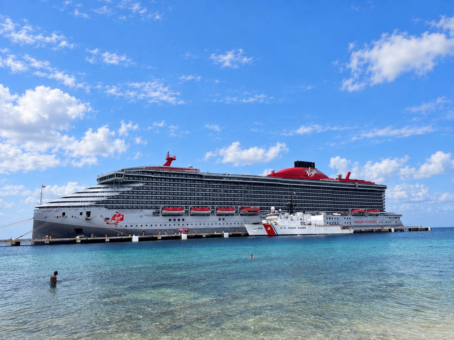 The Virgin Voyages Resilient Lady cruise ship docked in a Caribbean port beside a U.S. Coast Guard vessel.