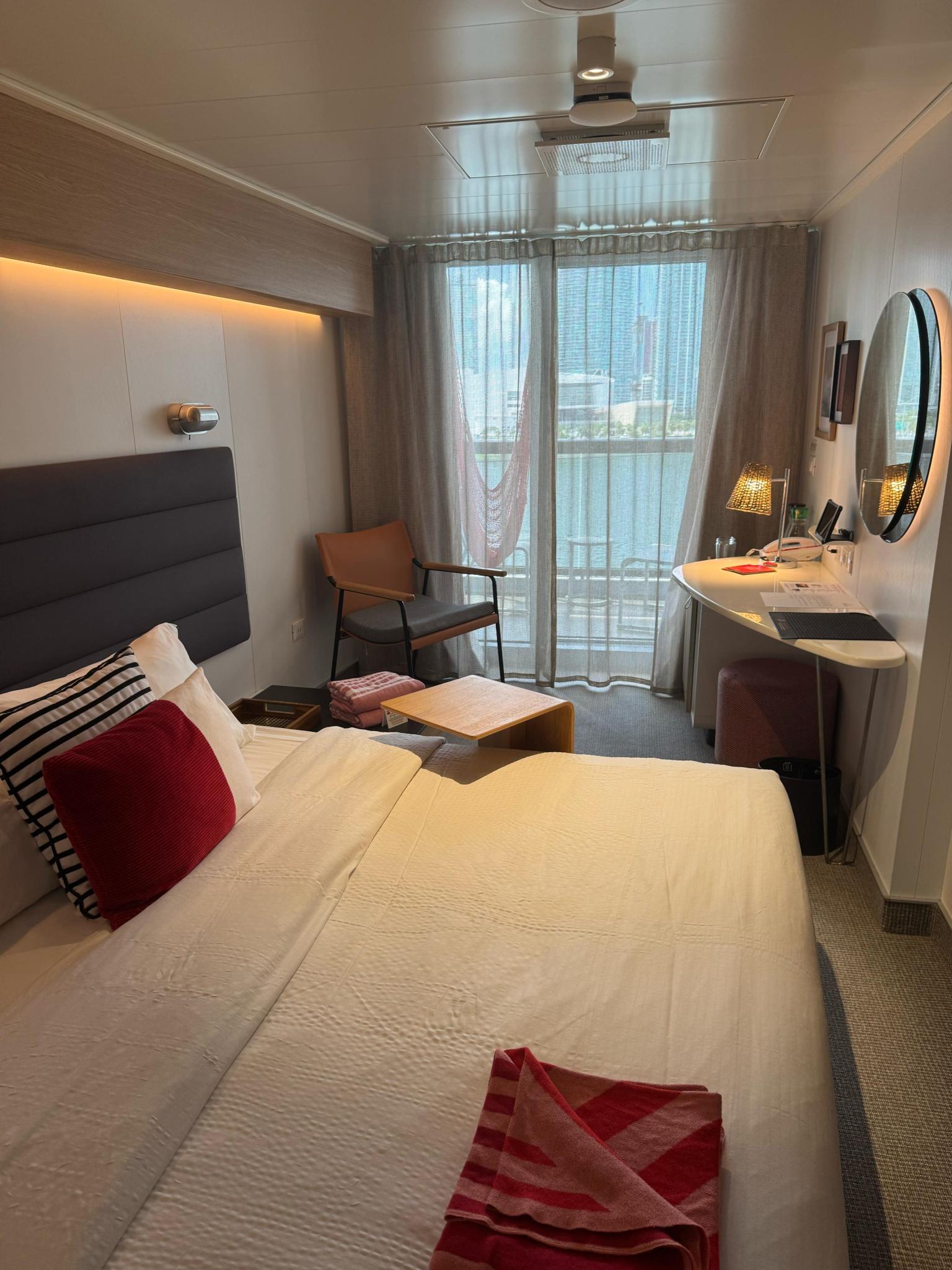A modern cruise ship cabin featuring a white bed, balcony, and city skyline view outside.