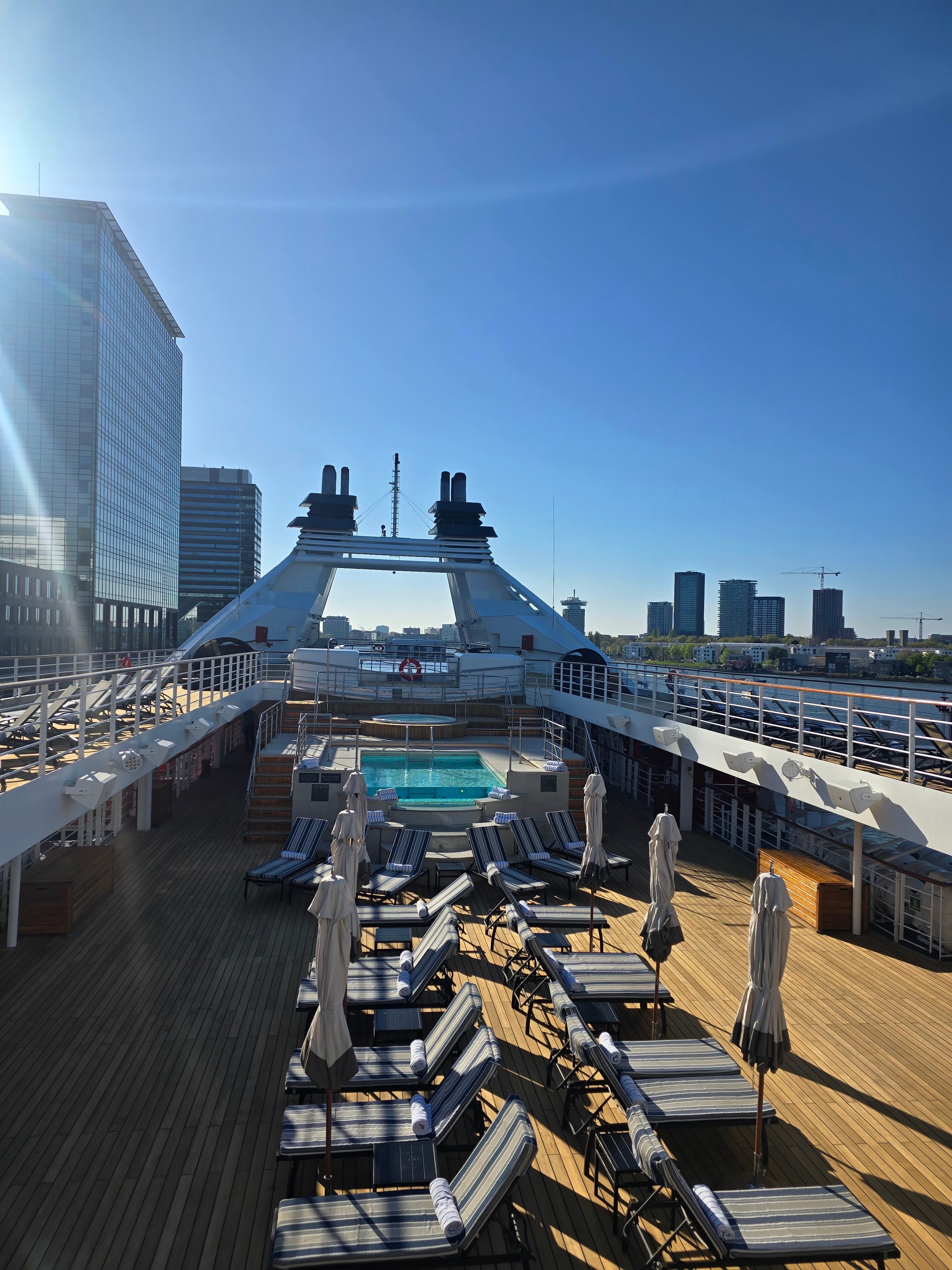 Aerial view of a cruise ship's pool deck with lounge chairs, umbrellas, and city skyline in background