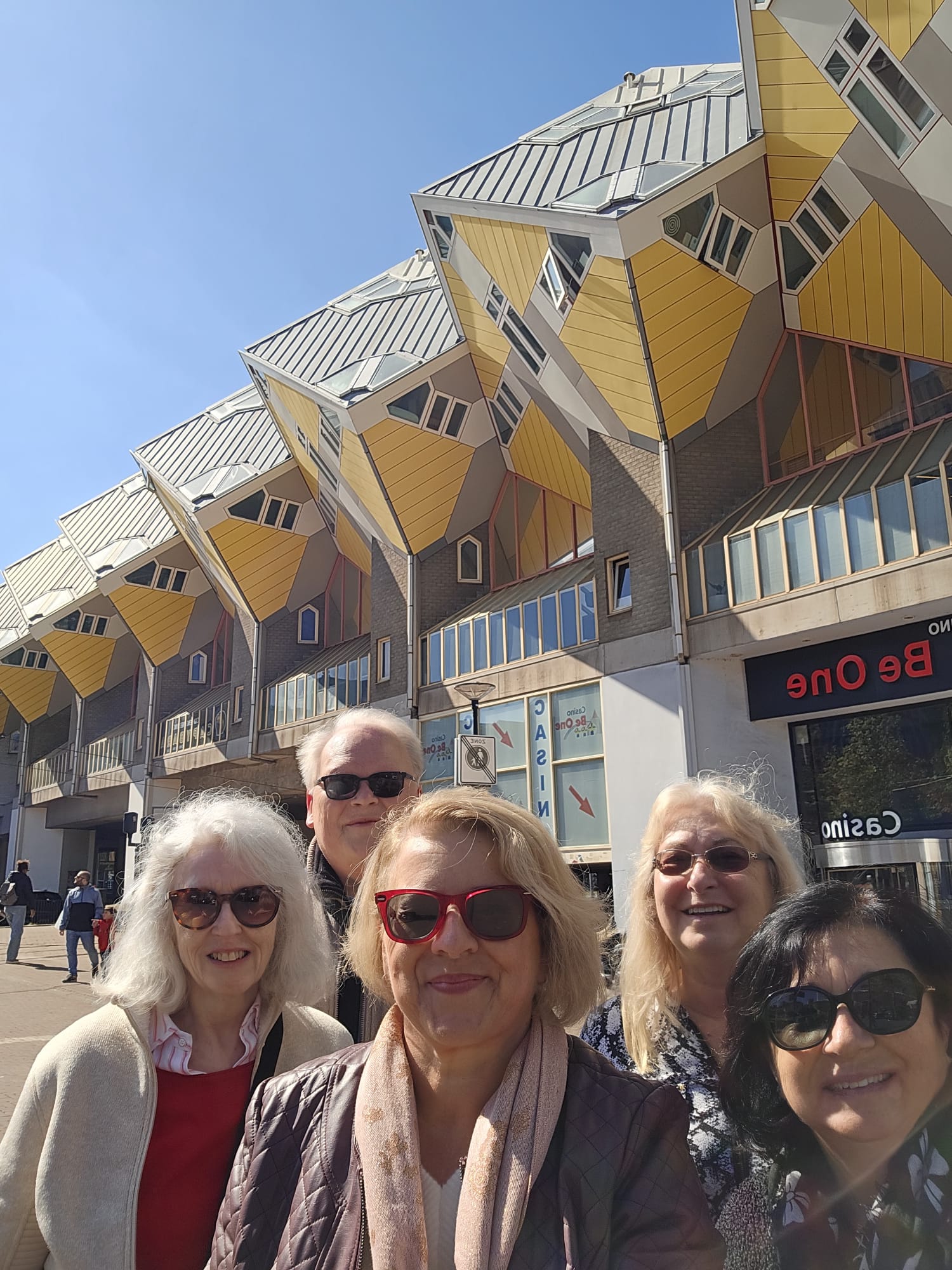 Group of five smiling women posing outdoors in front of distinctive yellow cube-shaped houses