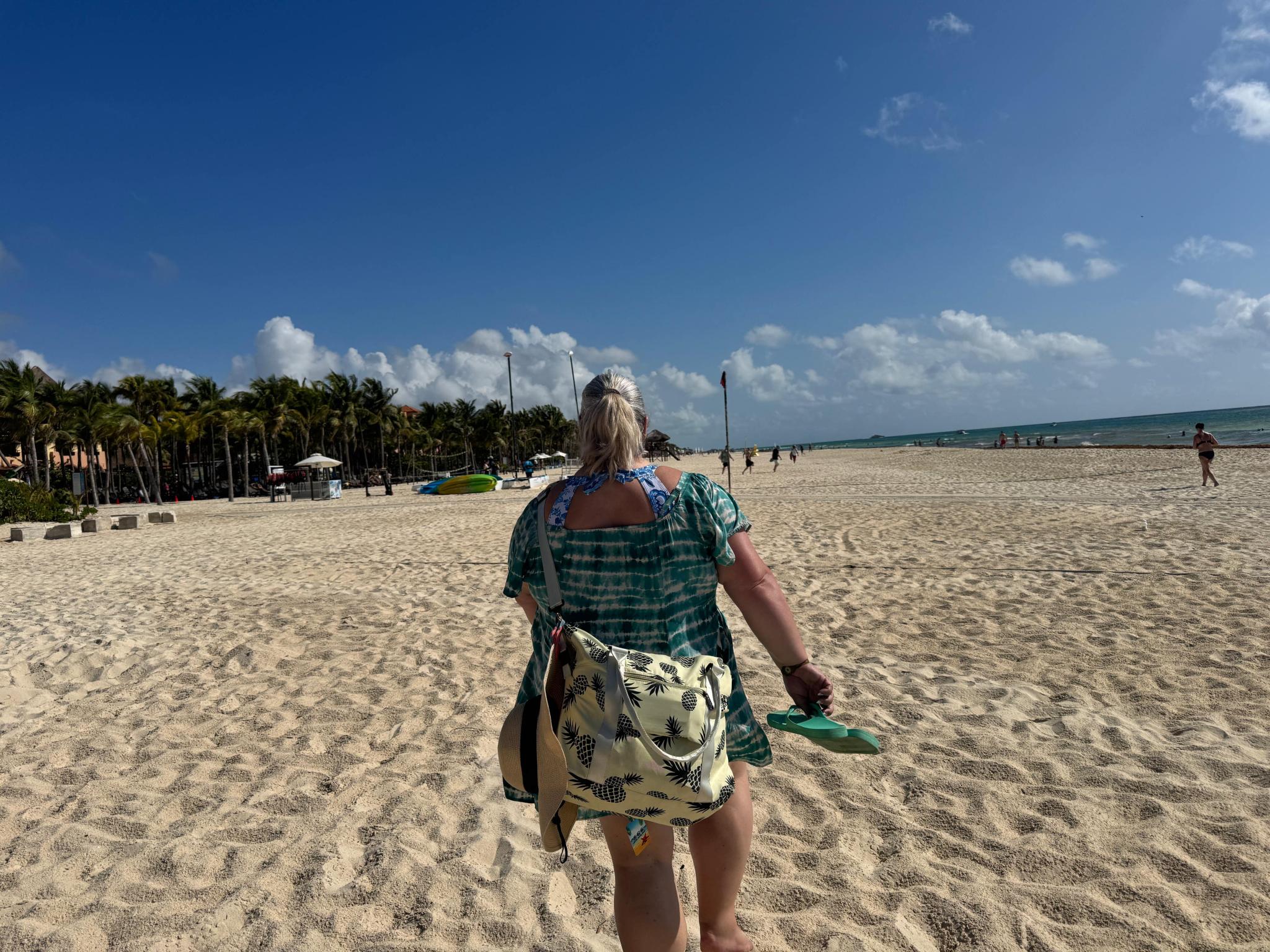 A woman walks barefoot across a wide white-sand Caribbean beach toward the ocean.