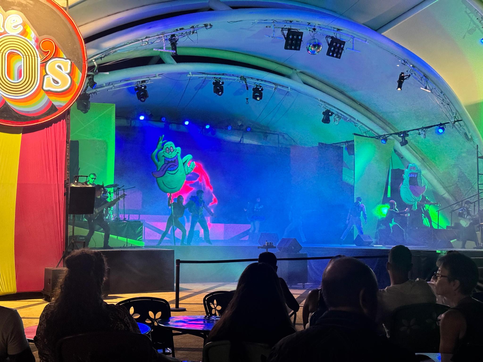 Audience watches a neon-lit 80s themed stage show featuring Ghostbusters Slimer imagery at a theme park.