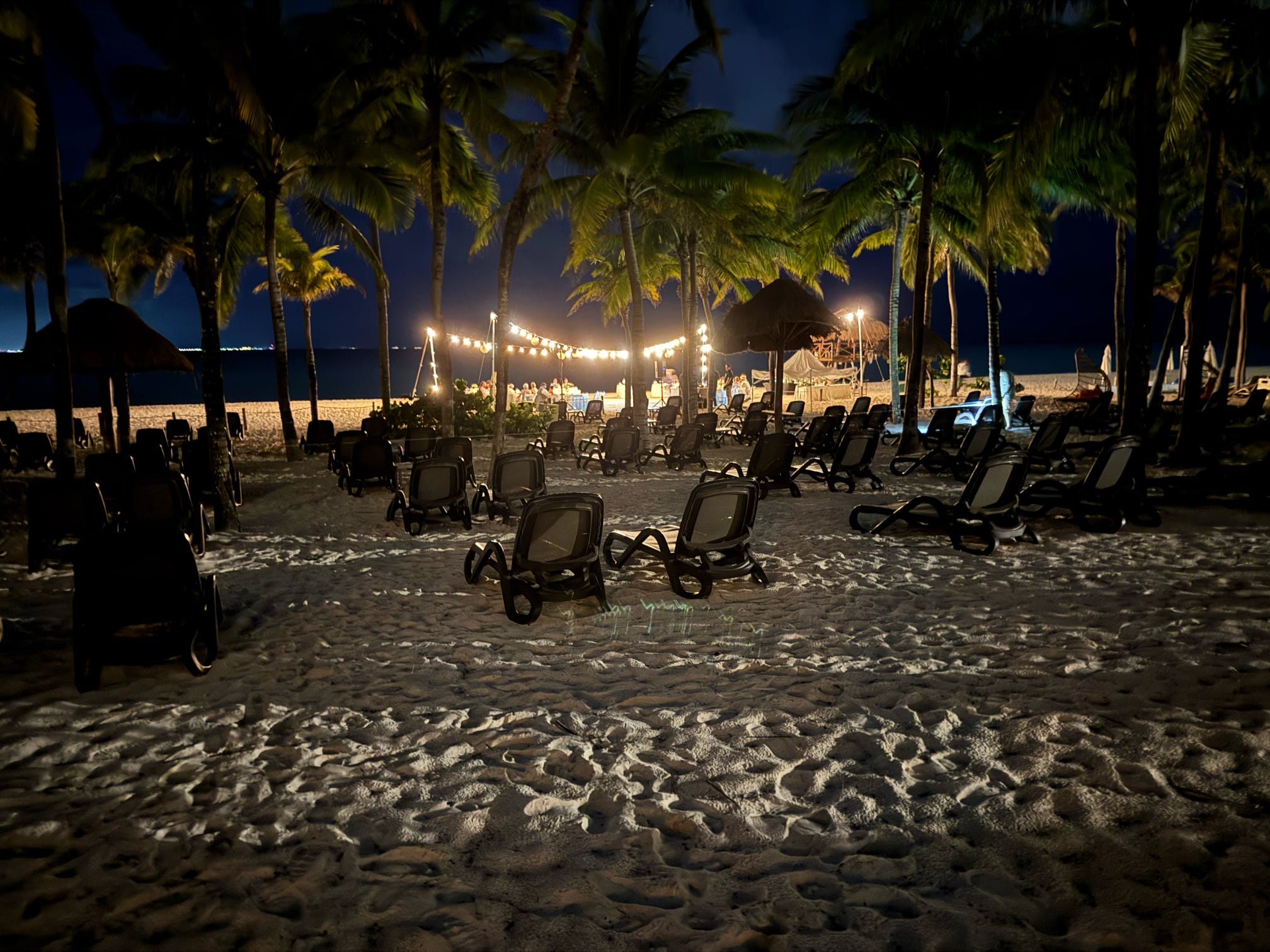 Empty beach loungers line a tropical resort shore at night, lit by warm string lights.