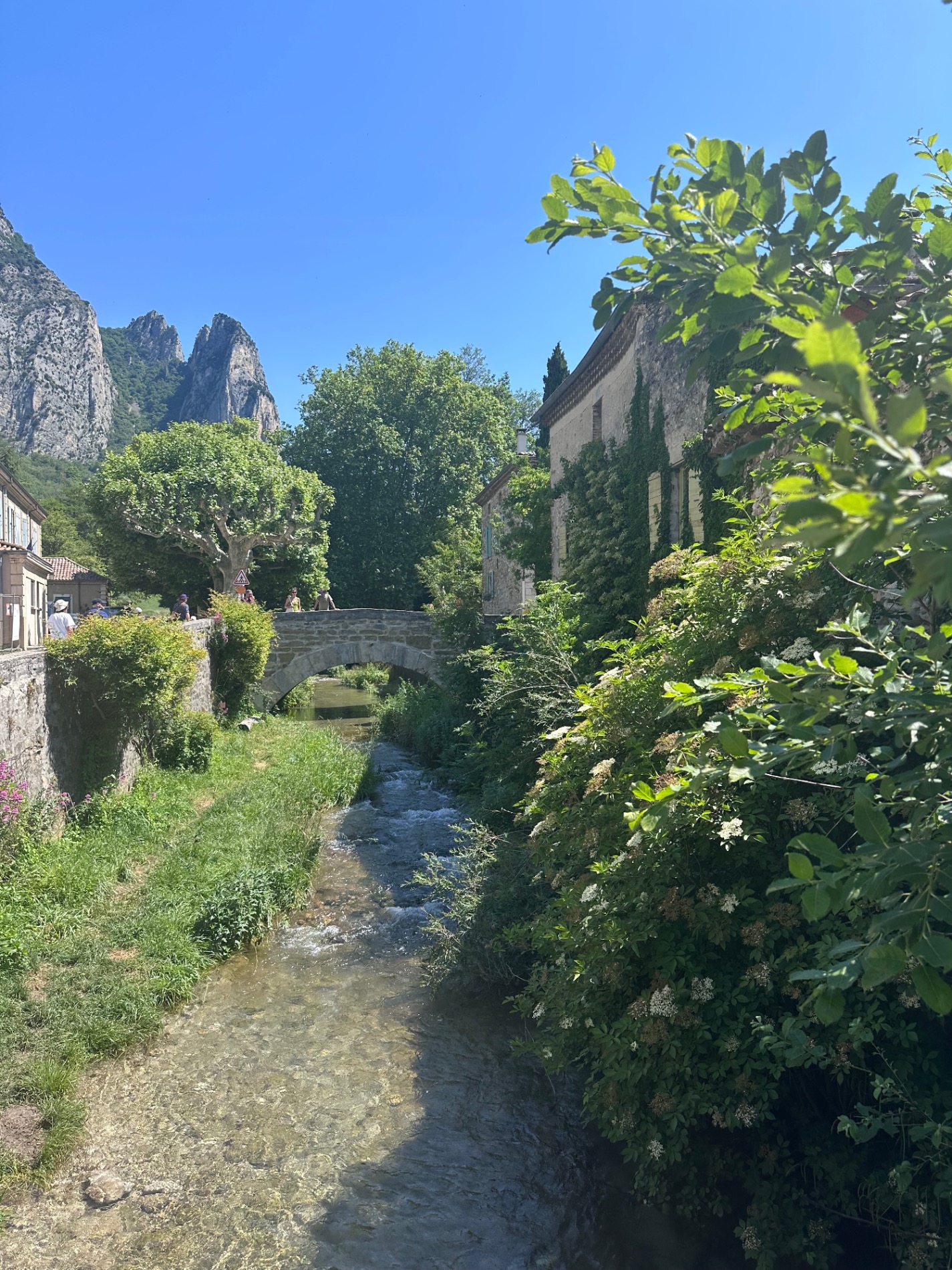 Stone buildings beside a flowing stream with lush green vegetation and dramatic rocky cliffs in background