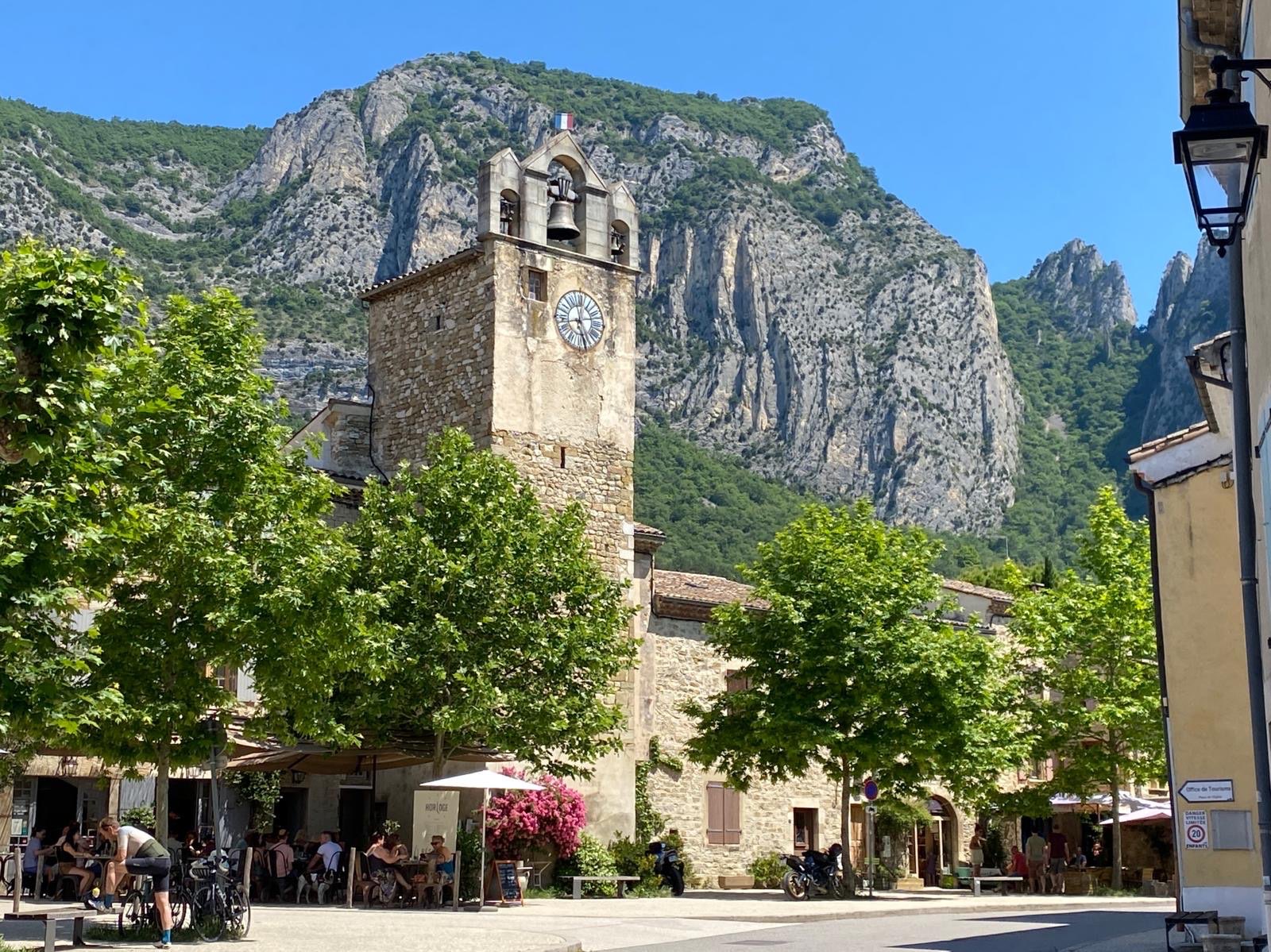 Clock tower of a stone church with multiple bells, surrounded by green trees and tall limestone rock formations