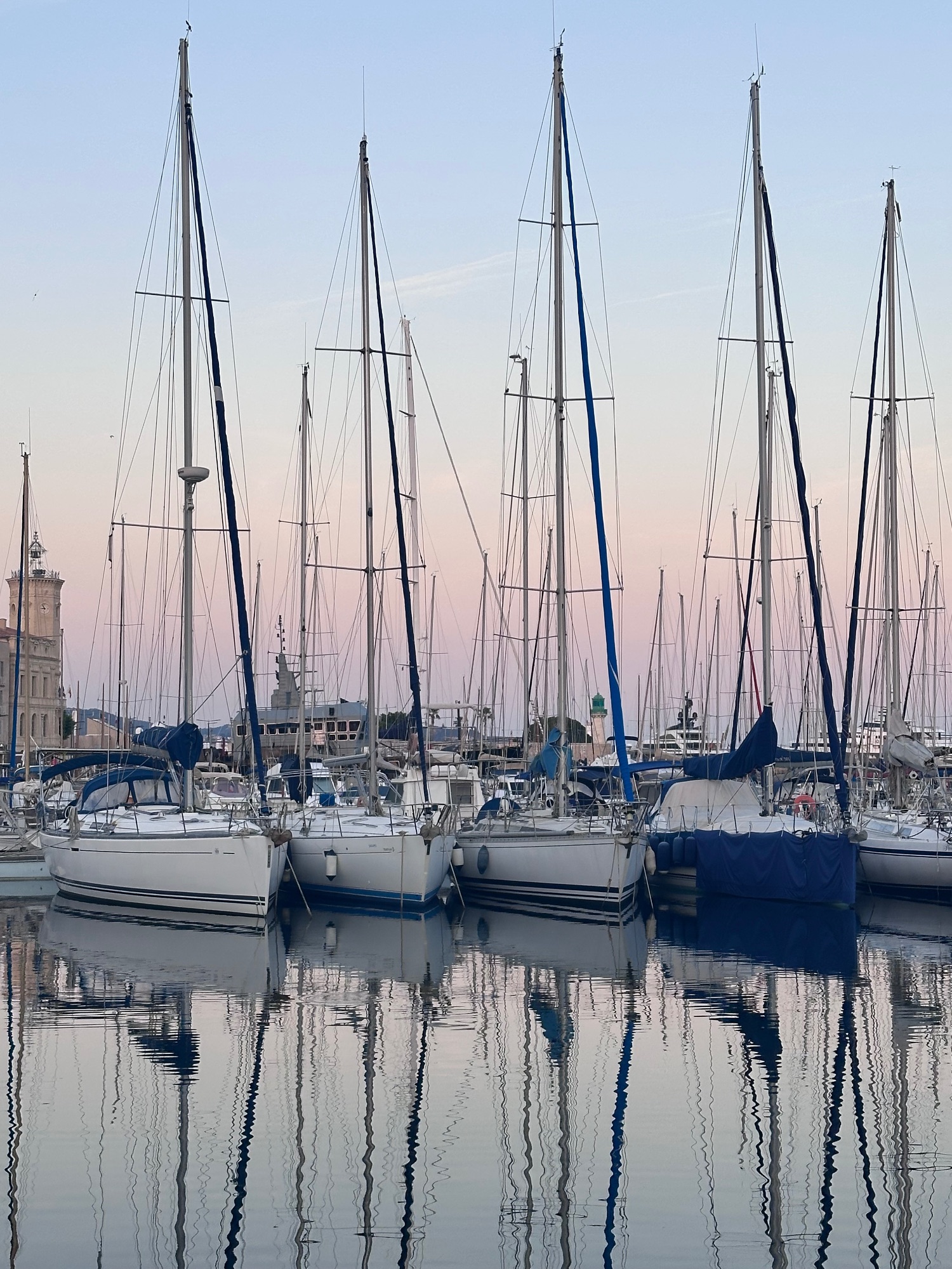 Sailboats with tall masts moored in calm water at dawn or dusk, their reflections visible on the glassy surface