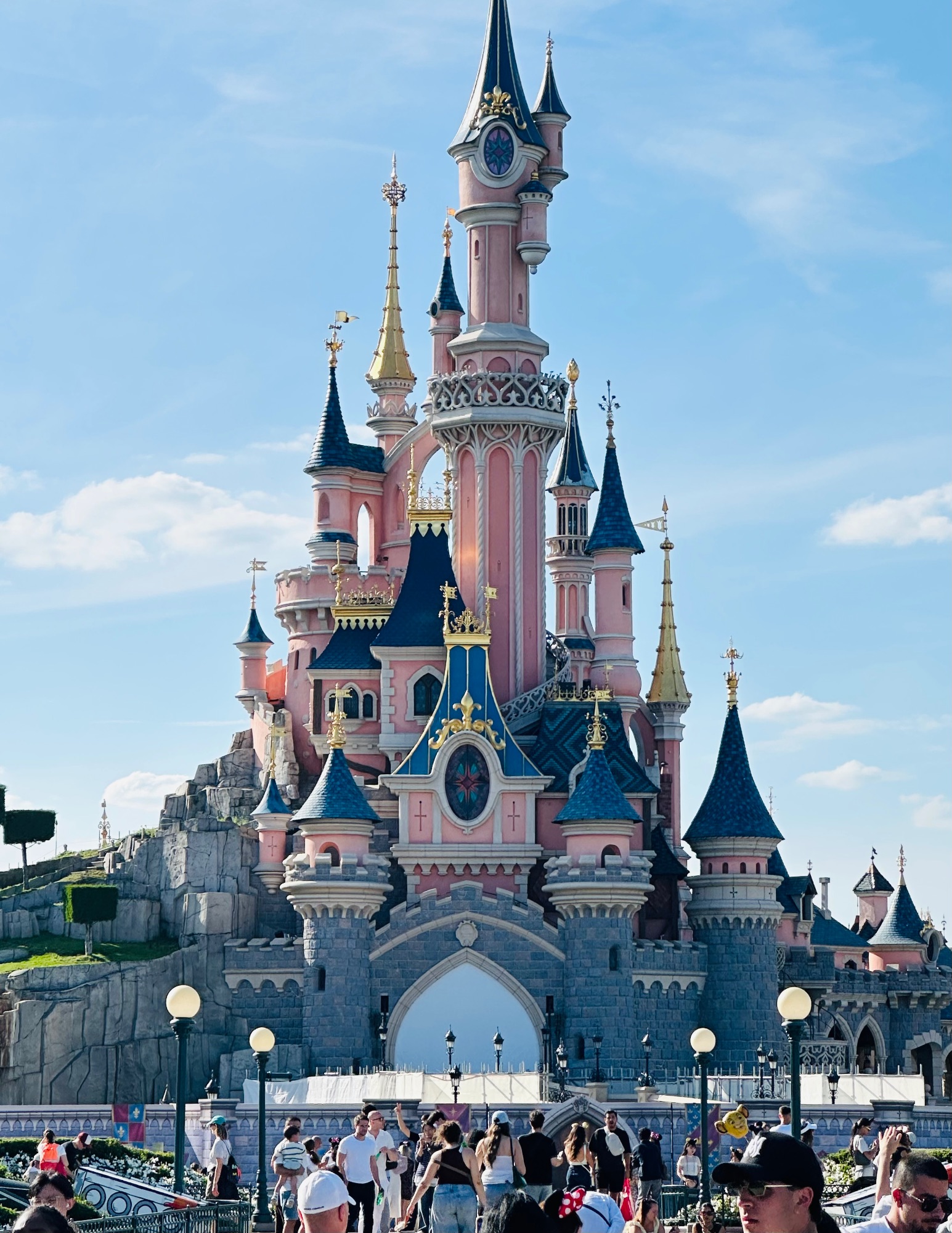 Pink and blue castle with multiple spires and towers against a blue sky, surrounded by crowds of visitors