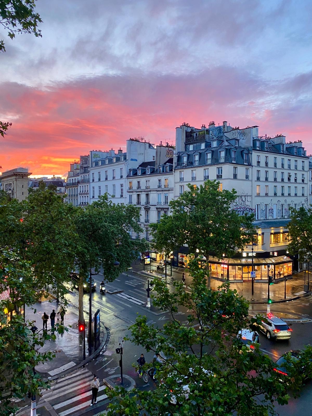 Parisian street at sunset with illuminated storefronts, classical buildings, and dramatic pink and orange sky