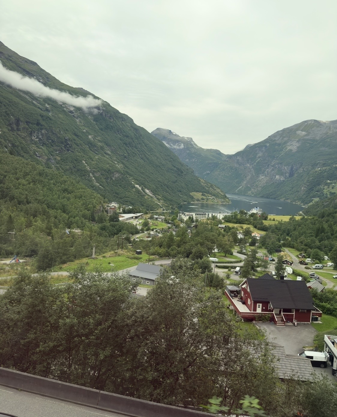 Aerial view of a Norwegian valley with a red house, forested slopes, and a deep blue fjord surrounded by mountains