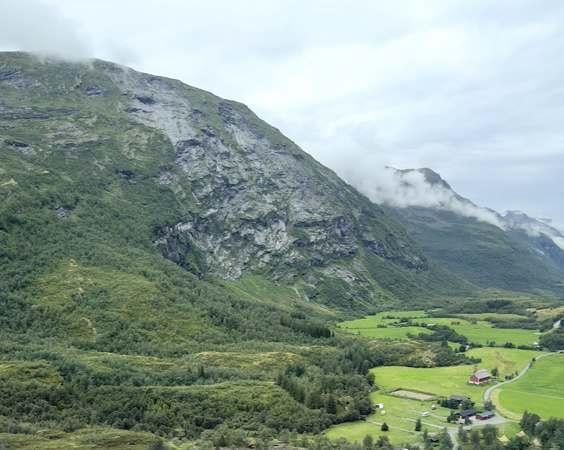 Aerial view of a cruise ship docked in a narrow fjord with red buildings, green fields, and forested mountains