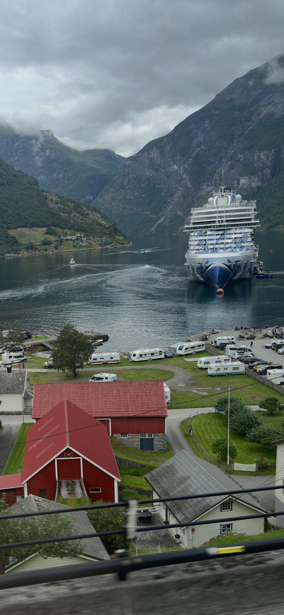 Cruise ship moored in a fjord with red farmhouse in foreground, green valley, and forested mountains rising steeply