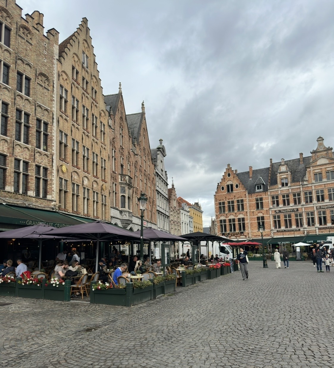 Medieval European town square with ornate brick buildings, market stalls with umbrellas, and outdoor dining areas