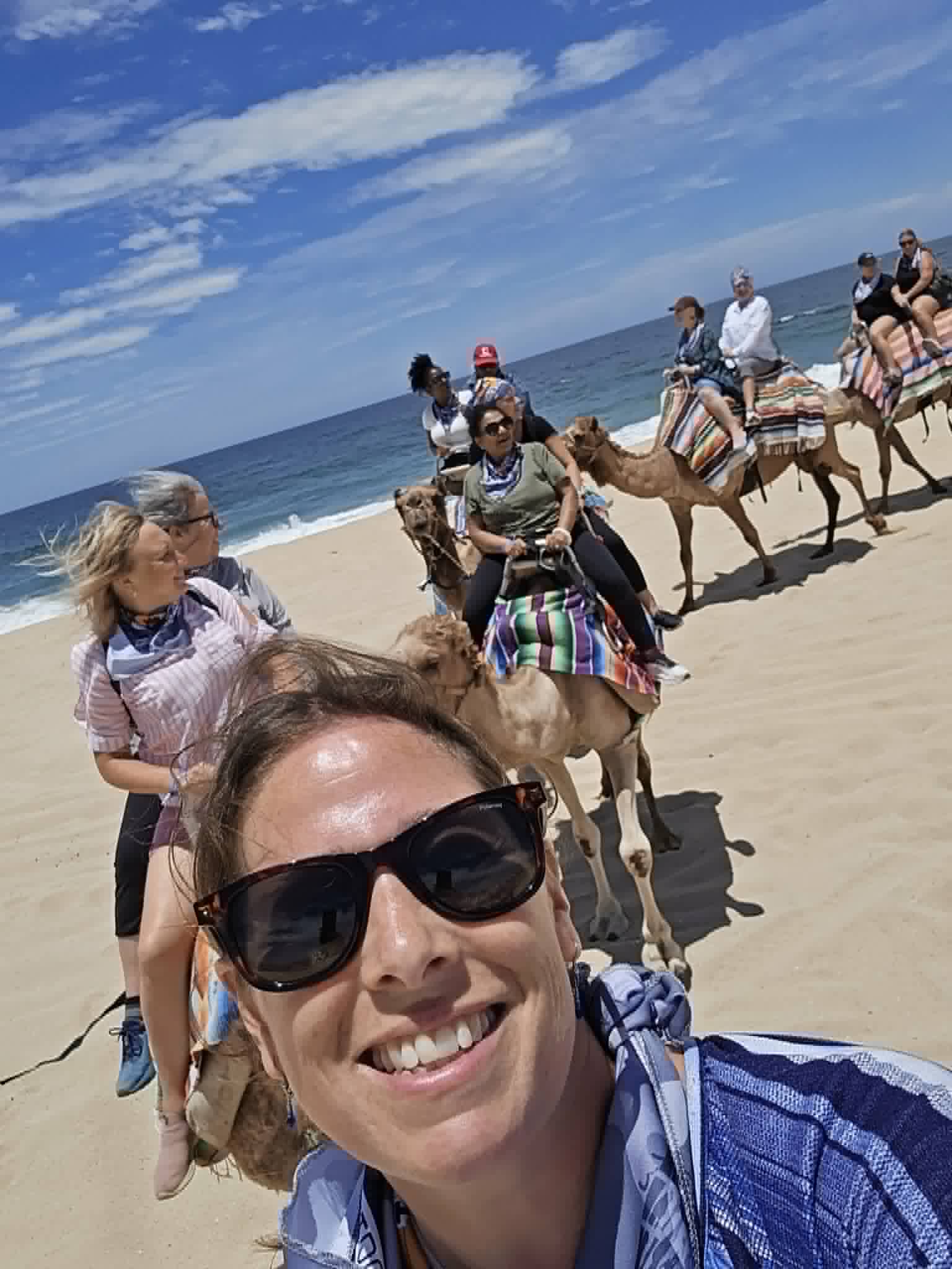 A woman in sunglasses rides an ATV on a beach with other riders and camels visible in the background