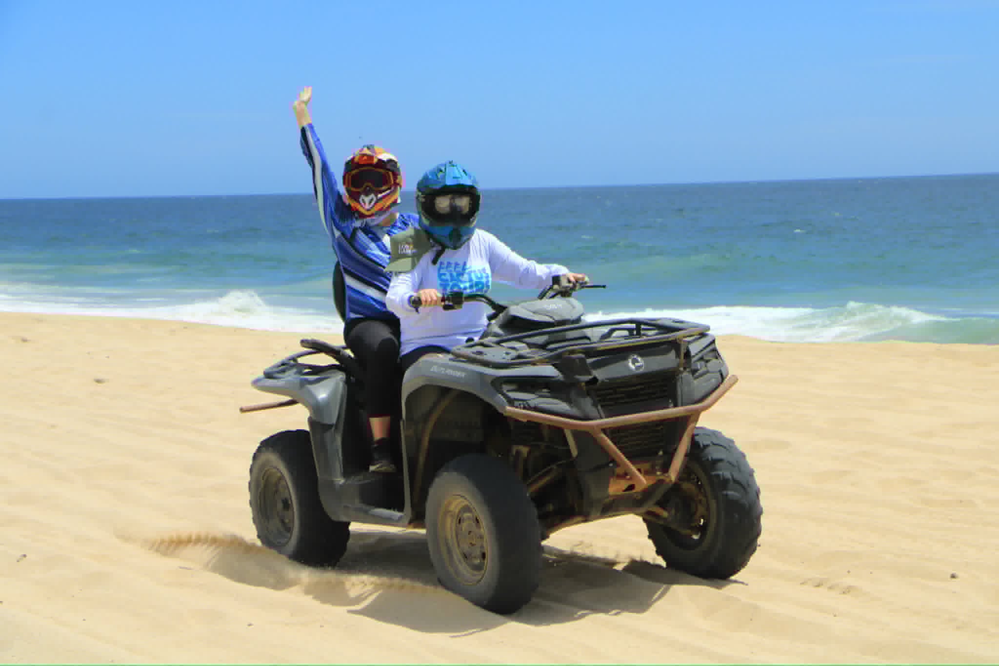 Two people in helmets celebrate on an ATV on a sandy beach with the ocean and blue sky behind them