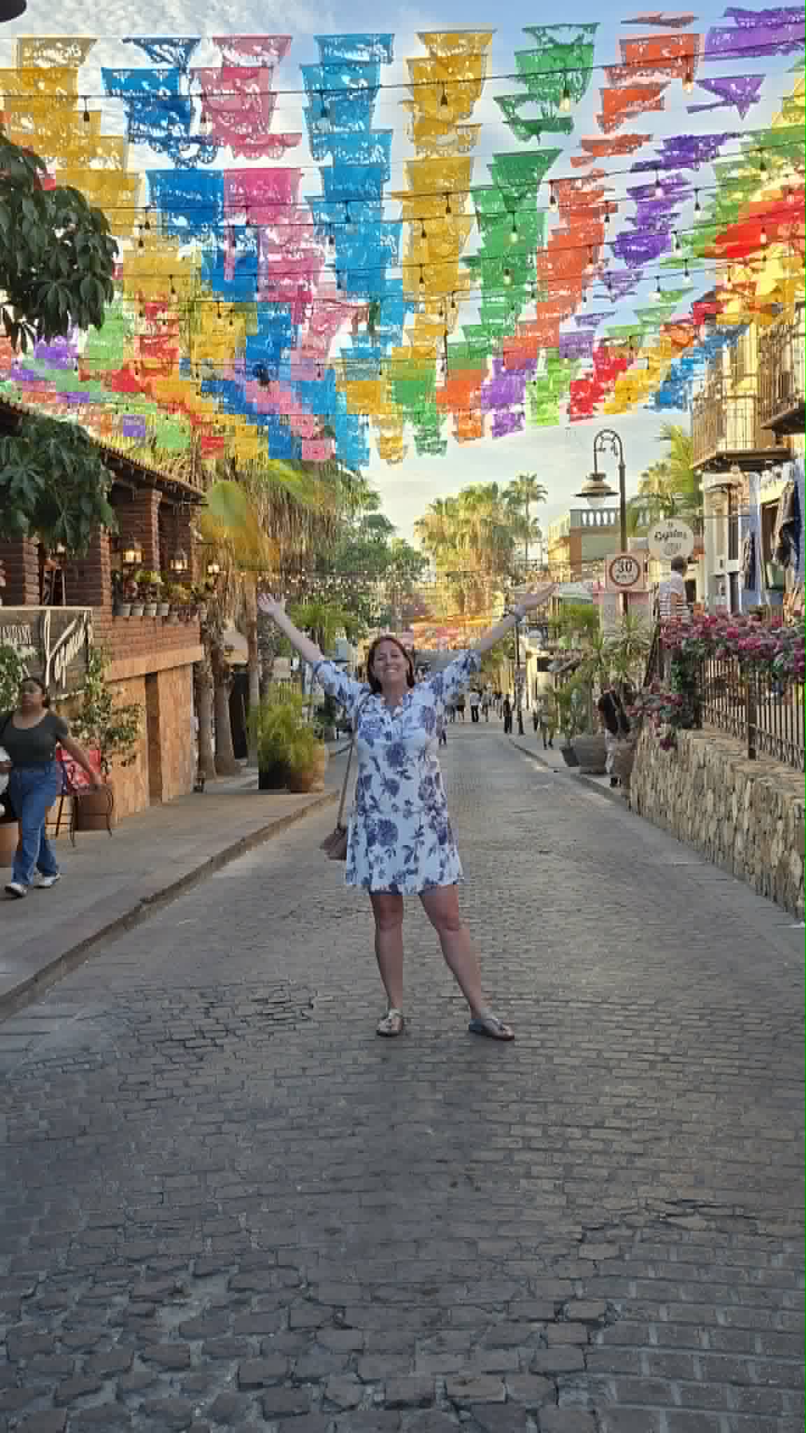 A woman stands under vibrant rainbow-colored papel picado decorations on a colonial-style street in Mexico