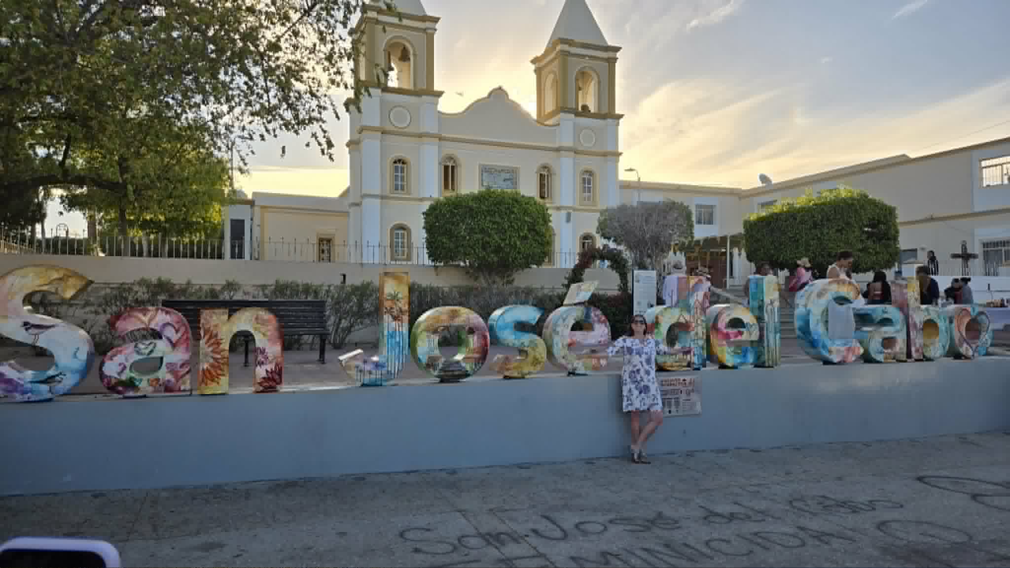 A woman poses at a large colorful mosaic sign reading 'San José del Cabo' in front of a white church