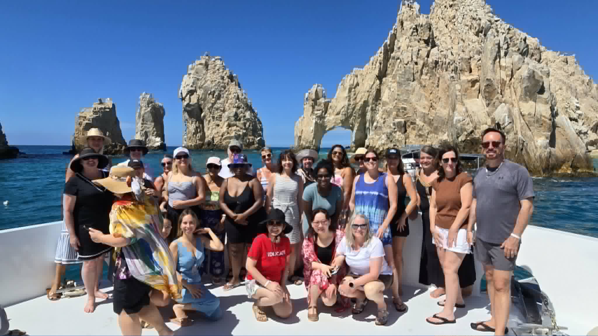 A group of tourists on a boat pose in front of dramatic rock formations and turquoise water at Cabo San Lucas