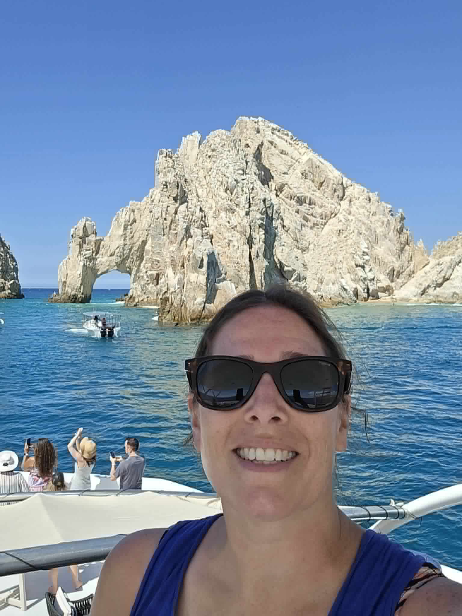 A woman in sunglasses takes a selfie on a boat with distinctive rock arch formations visible in the ocean behind her