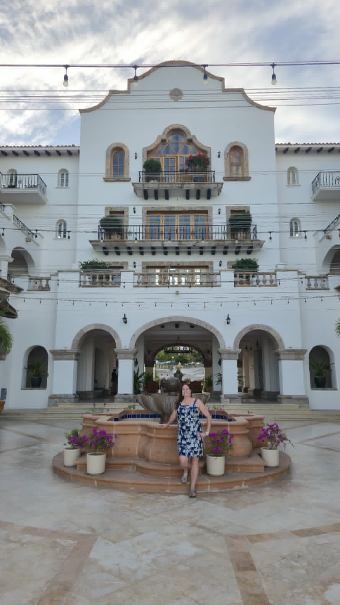 A woman stands in front of a white Spanish colonial building with arched doorways and flowering vines