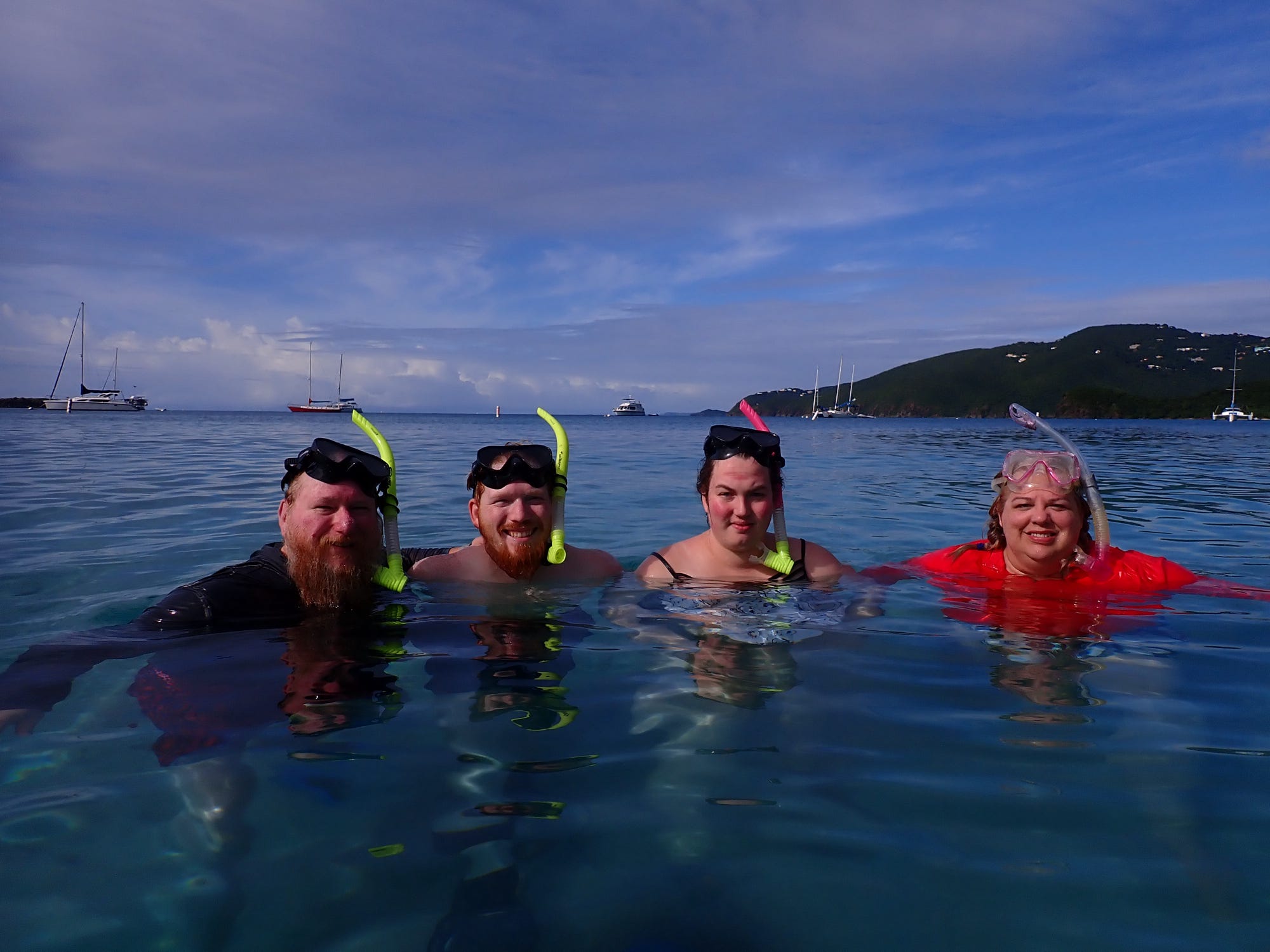 Four smiling snorkelers float in clear Caribbean waters with sailboats and a green island behind them.