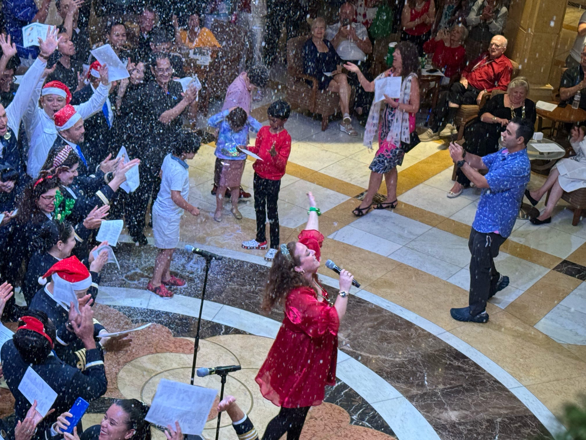 A performer in red leads a joyful Christmas carol sing-along with artificial snow in a cruise ship atrium.