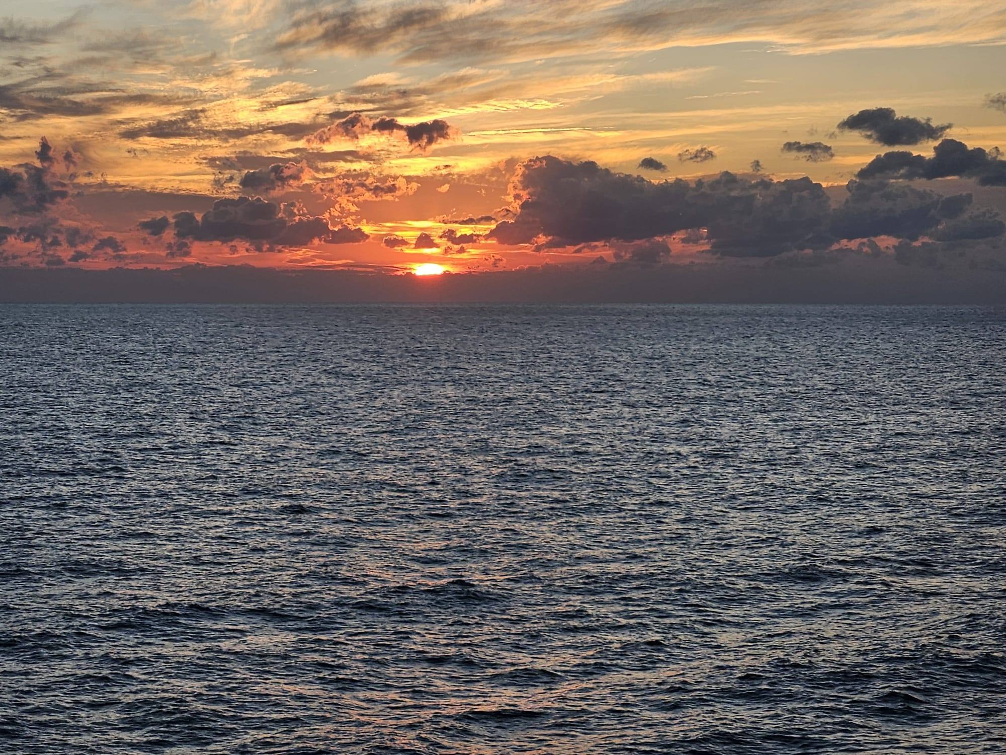 A dramatic fiery sunset blazes over a vast, gently rippling open ocean with moody clouds.
