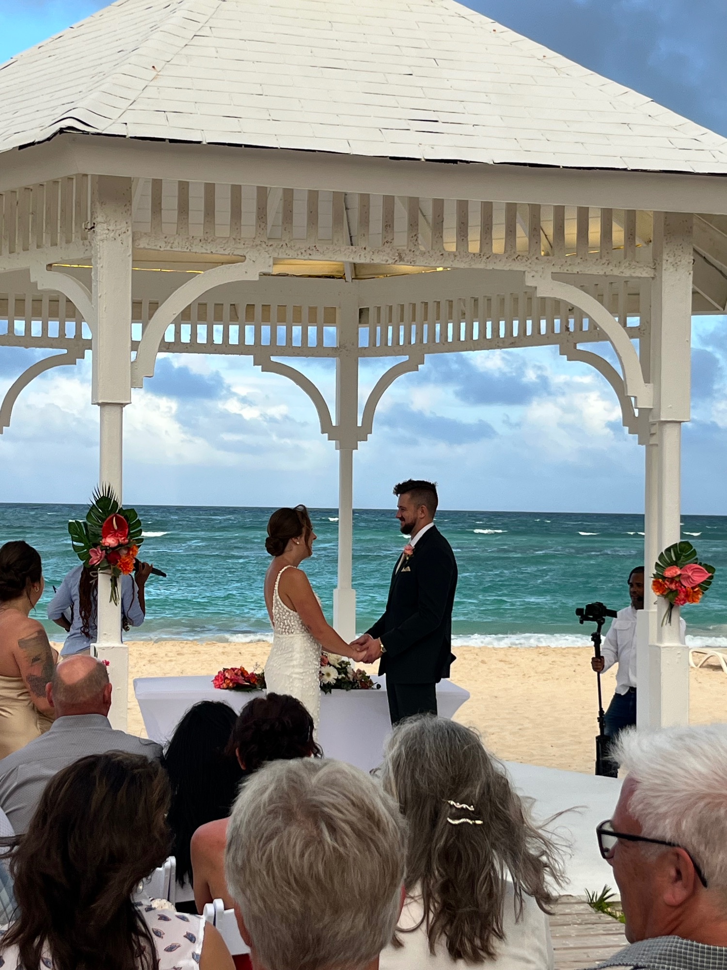 A bride and groom exchange vows under a white gazebo on a beach in Punta Cana with turquoise ocean and guests visible