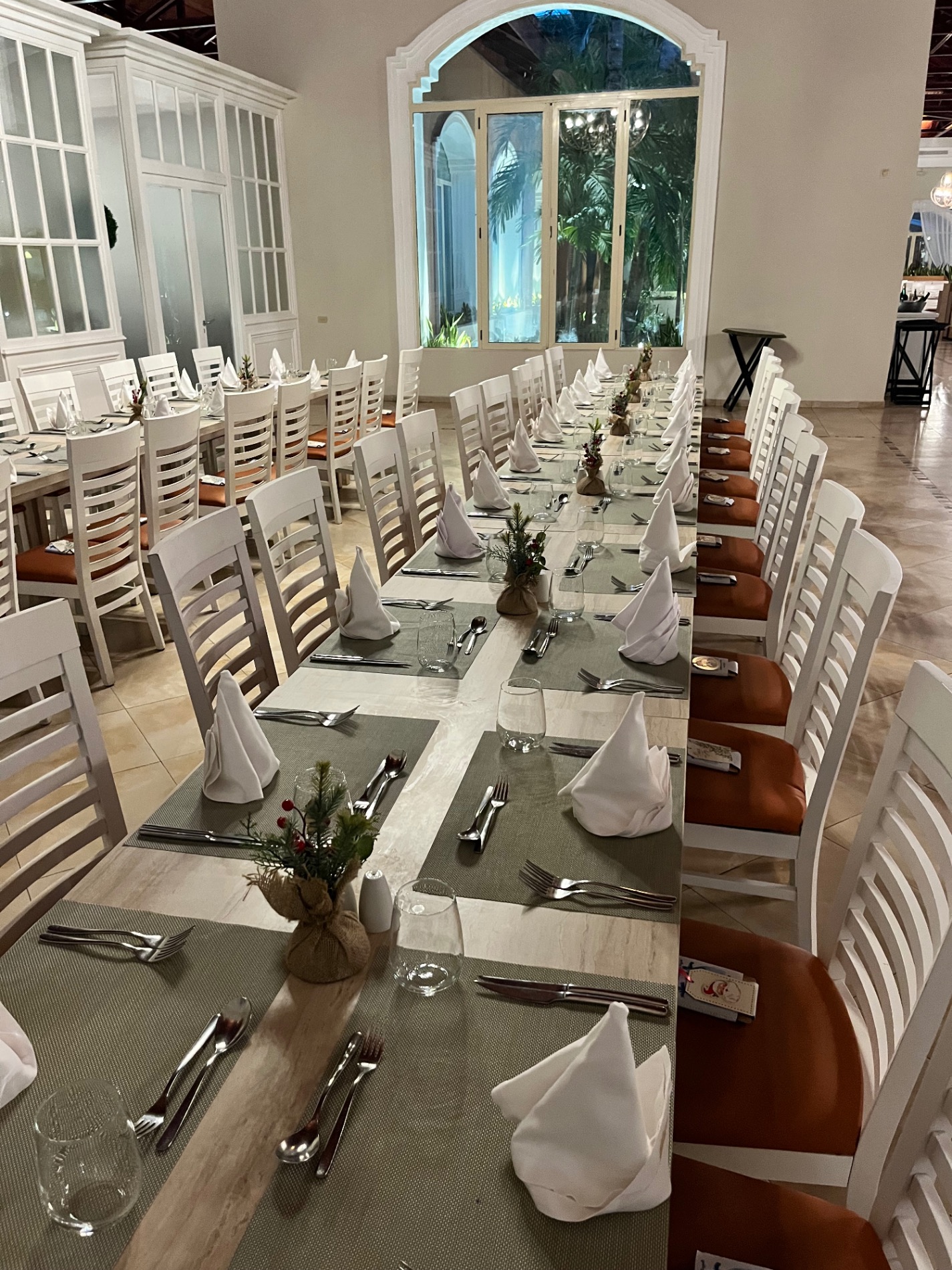 An elegant indoor reception hall with long white-clothed tables, white chairs, and tropical floral centerpieces