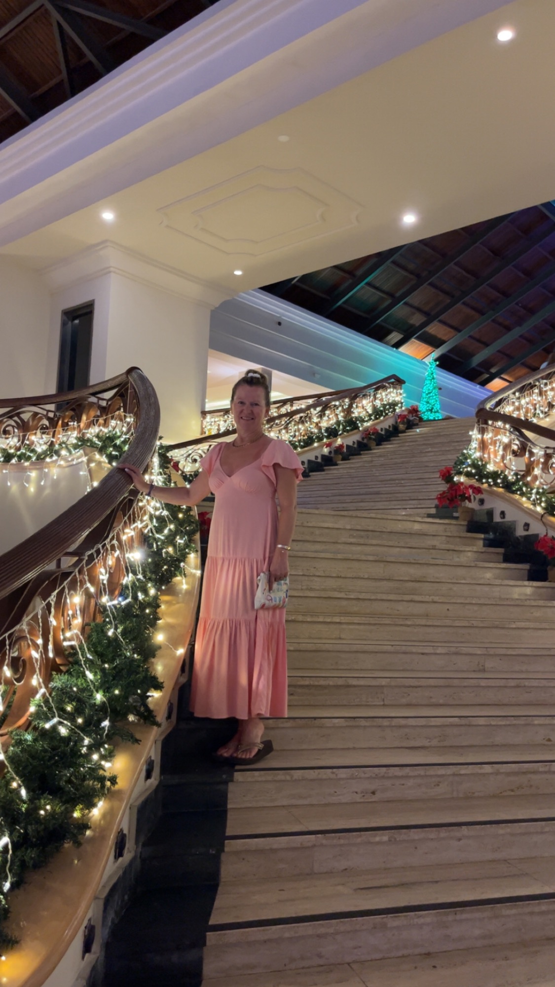 A woman in a pink dress stands on a grand staircase decorated with garland and white lights at an upscale resort