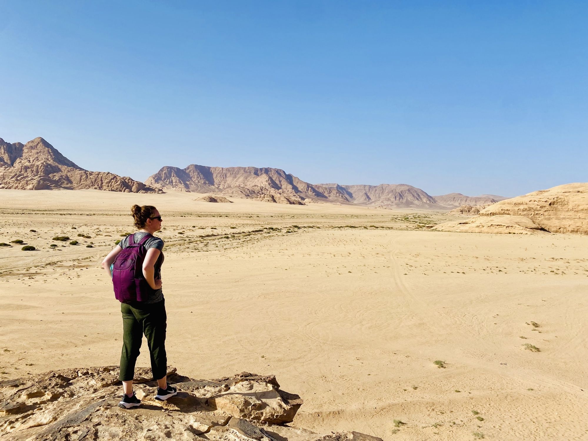 Autumn Fiske stands in a vast desert landscape with rocky mountains in the distance under clear blue sky in Jordan
