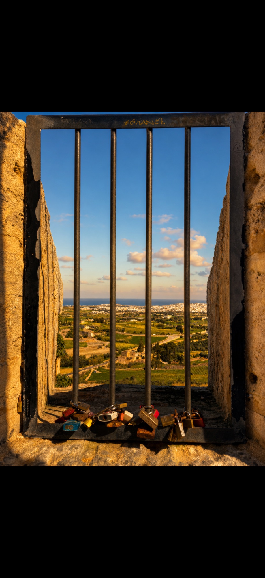 Barred window opening onto expansive countryside, Mediterranean sea, and scattered villages under blue sky