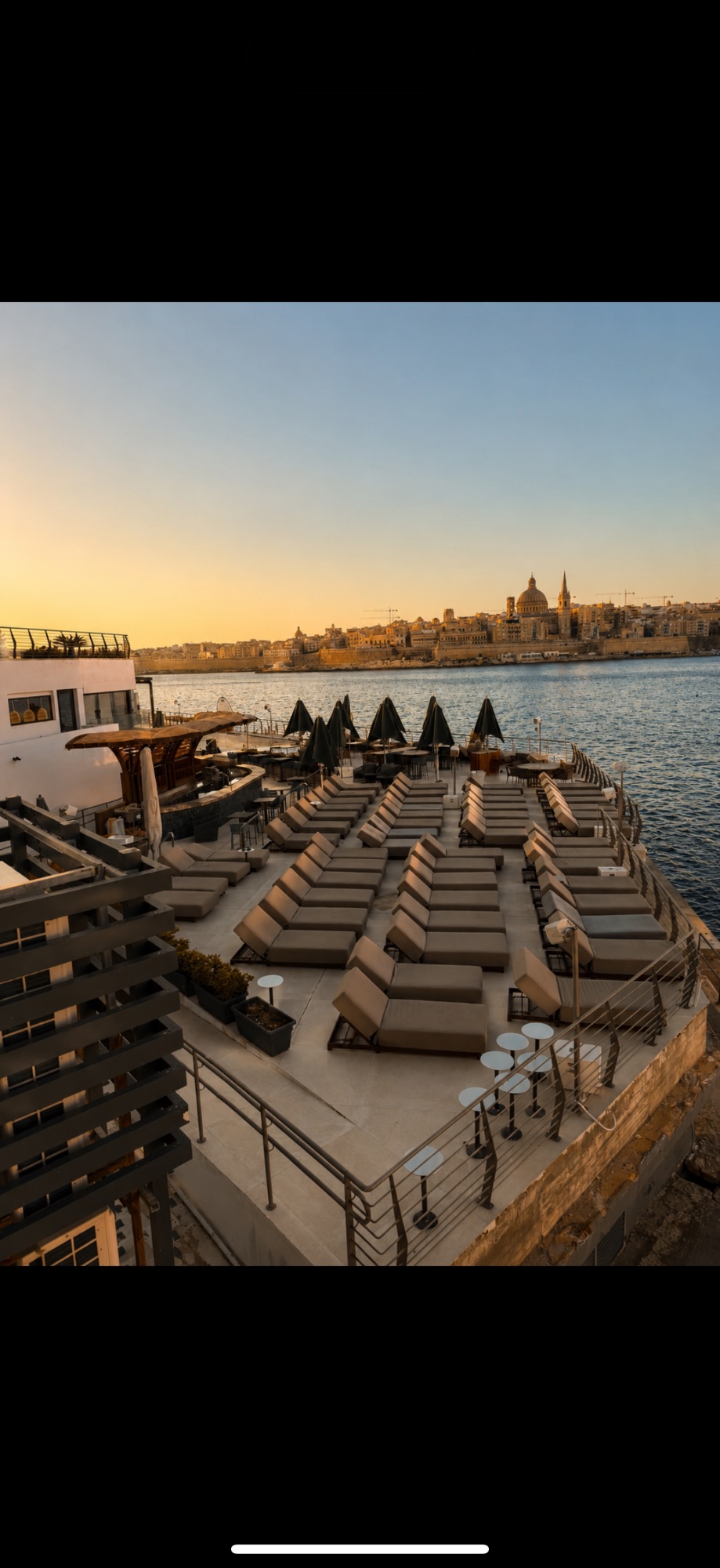 Sunbed lounge area on a waterfront deck with Valletta's historic architecture glowing across the harbor at sunset