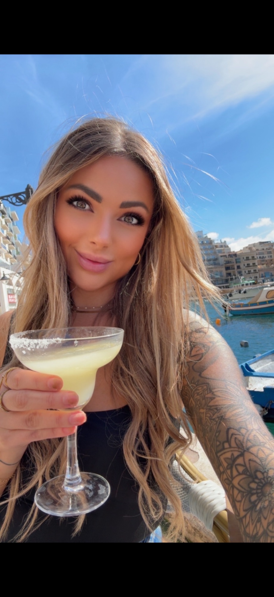 Bre Davis holding a cocktail at a Maltese harbor, with traditional fishing boats and waterfront buildings behind her