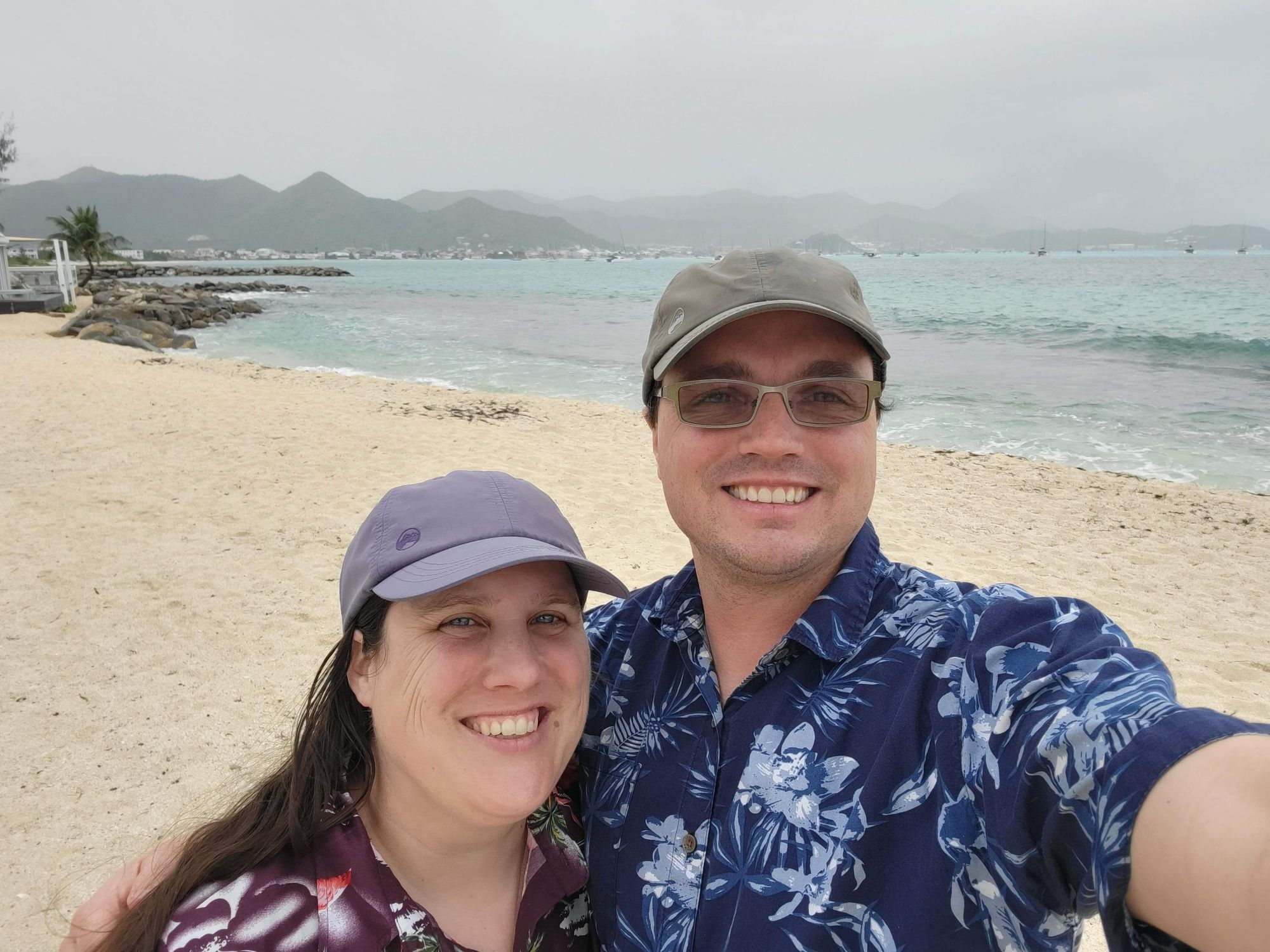 Couple smiling at camera on a sandy beach with green hills and anchored boats in the background