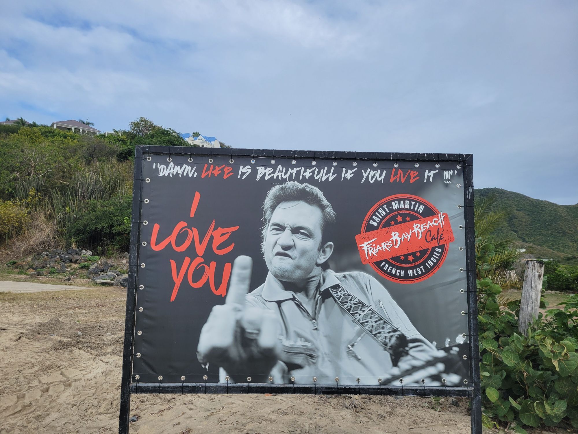 Billboard at Friars Bay Beach Café featuring a man's face with text 'Damn, life is beautiful if you live it' and 'I love you'
