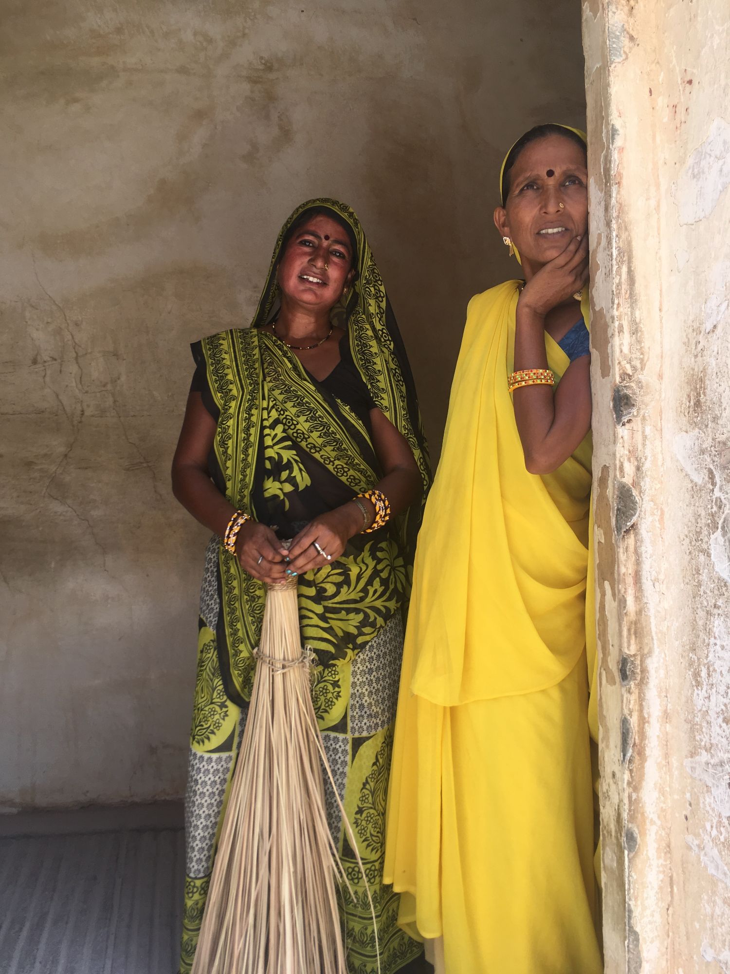 Two Indian women in traditional dress with decorative headscarves and bindis smile at the camera in a simple room